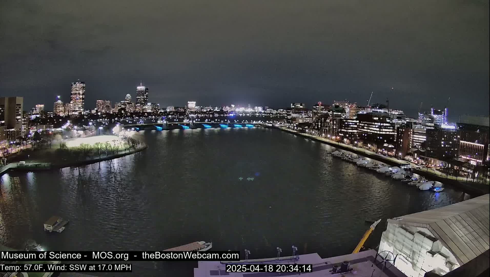 A nighttime view of a city skyline across a dark body of water, with a bridge illuminated in blue, under a dark, overcast sky.