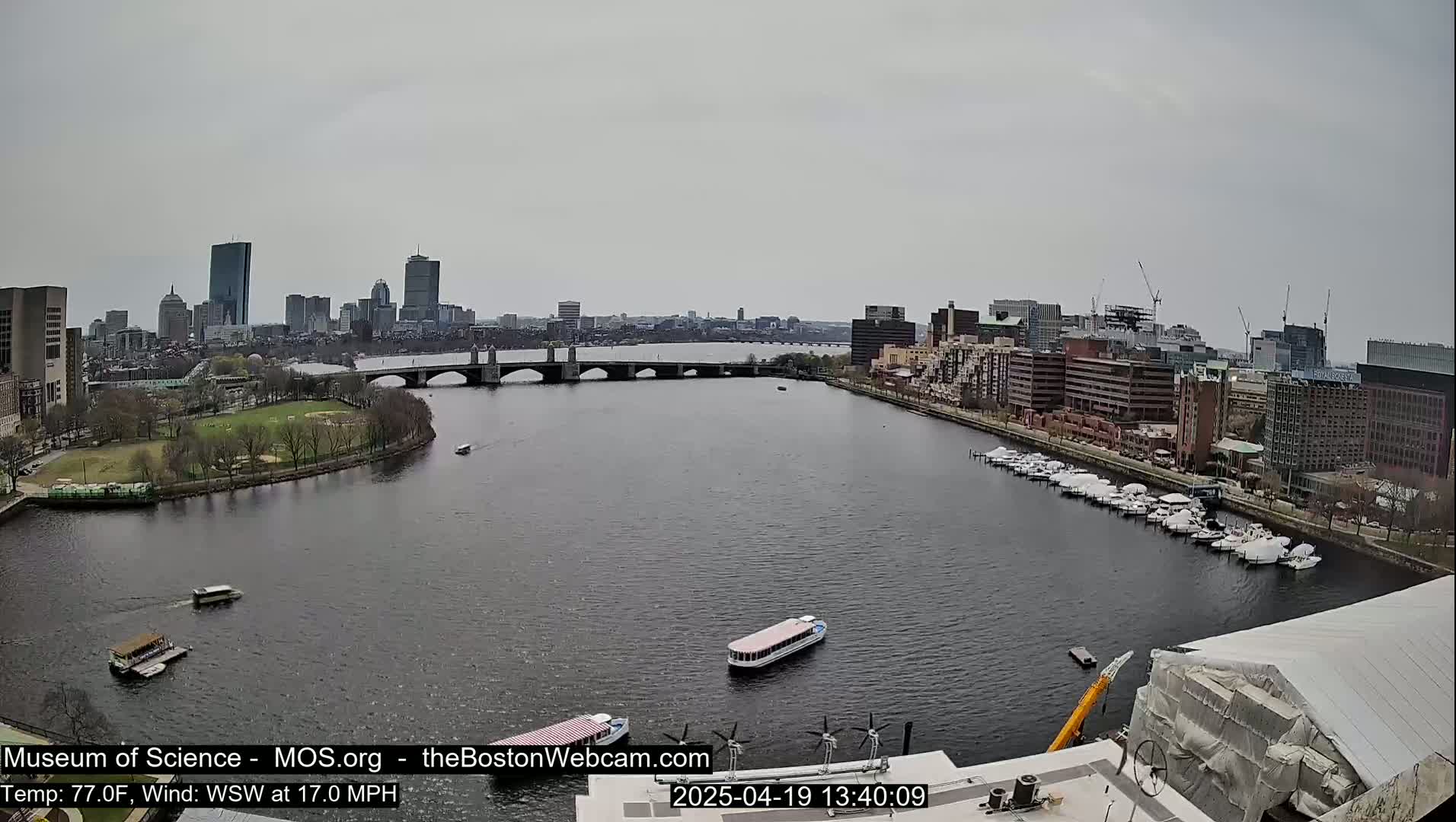 A city skyline under an overcast sky is viewed from across a river, with boats on the water and a bridge in the middle ground.