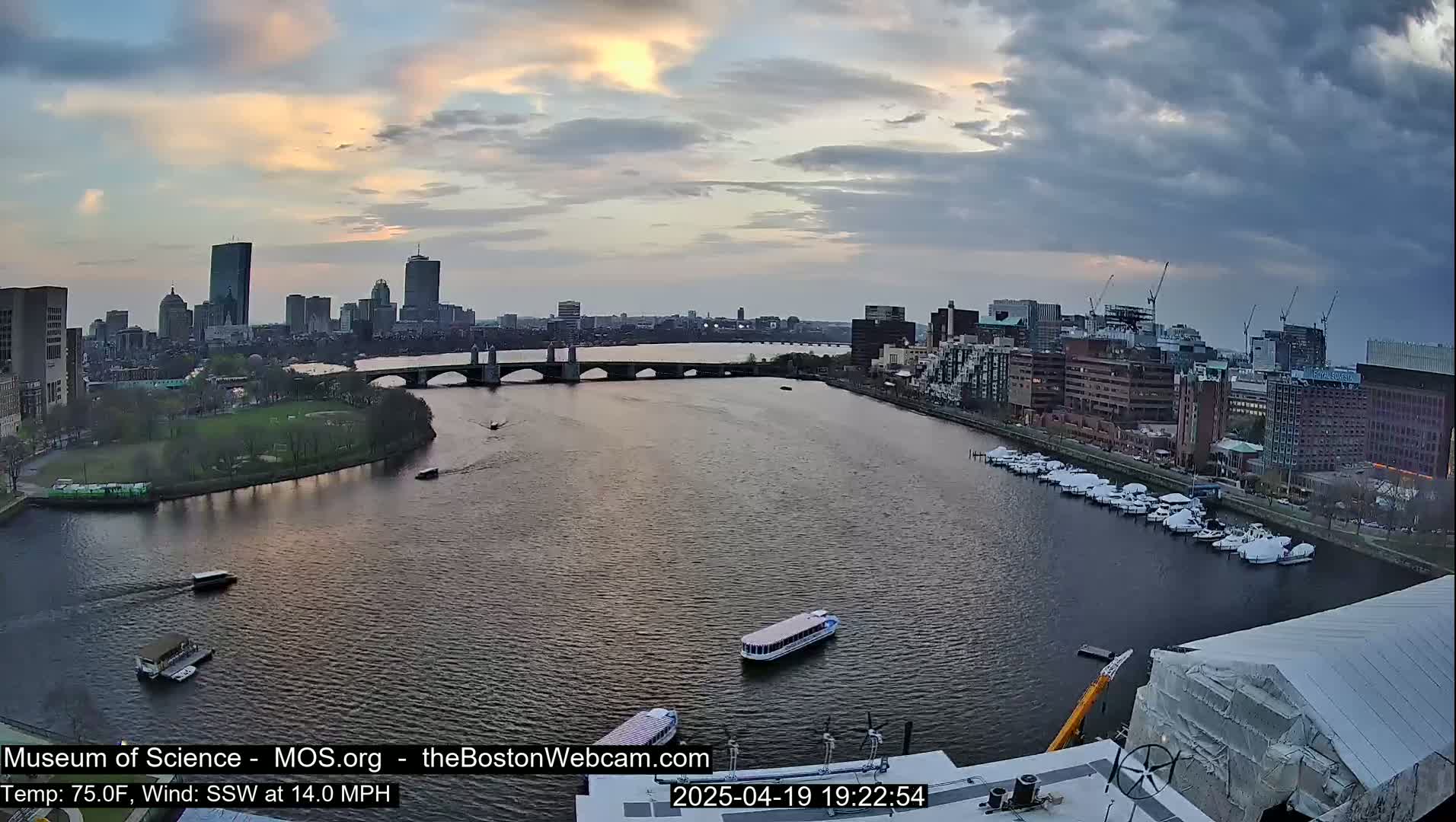 A wide, high-angle view shows a city skyline across a river at sunset under partly cloudy skies, with several boats on the water and a bridge in the middle ground.