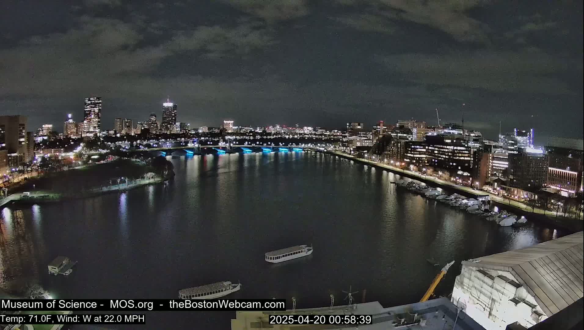 A nighttime cityscape across a dark river, with city lights reflecting on the water under a cloudy sky, and boats moored along the shore.
