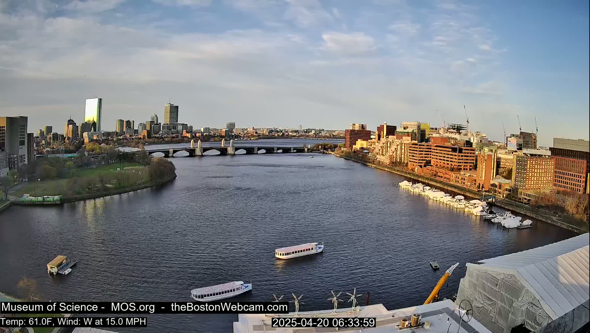 A city skyline is visible across a calm river under a partly cloudy sky, with several boats on the water and a bridge in the mid-ground.
