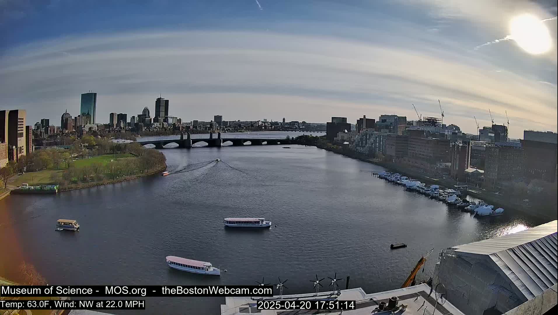 A sunny cityscape with a river, bridges, boats, and a park is viewed from a high vantage point.