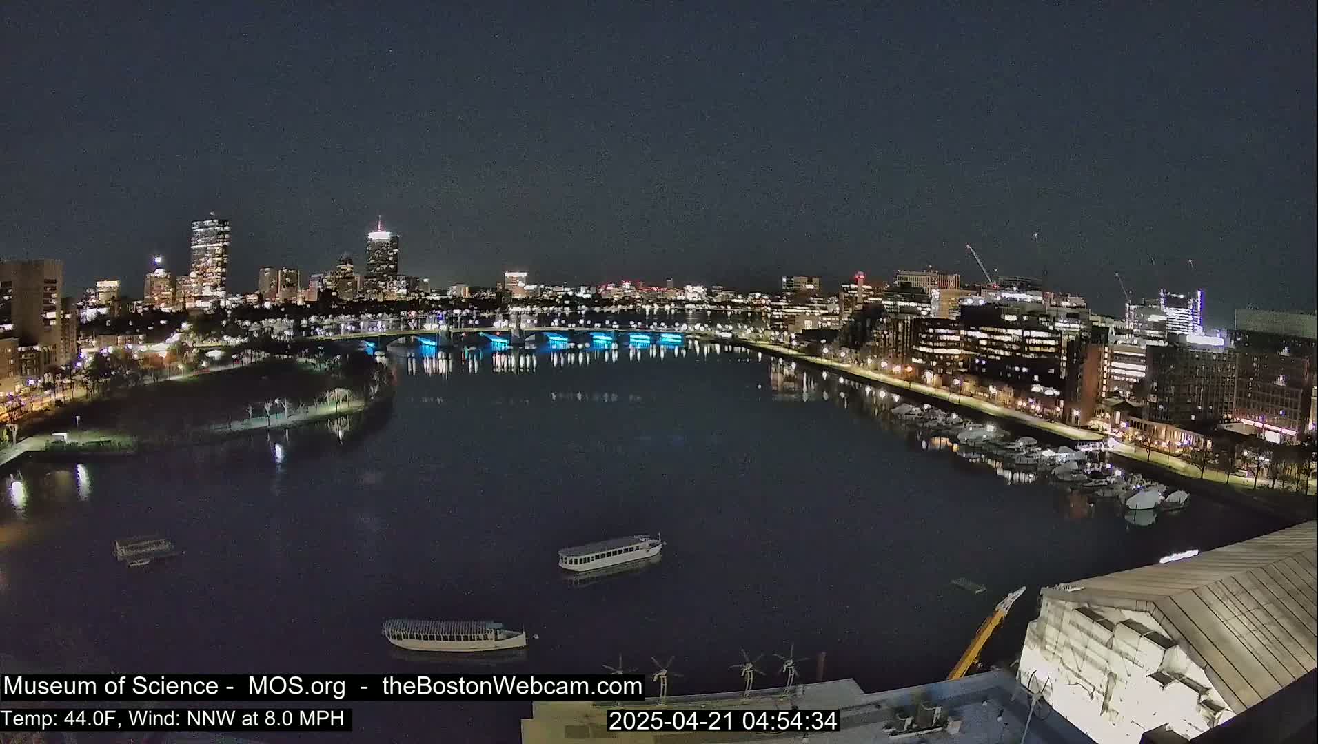 A nighttime view of a city skyline across a calm river, with boats docked along the shore and a few small vessels on the water under a clear, dark sky.