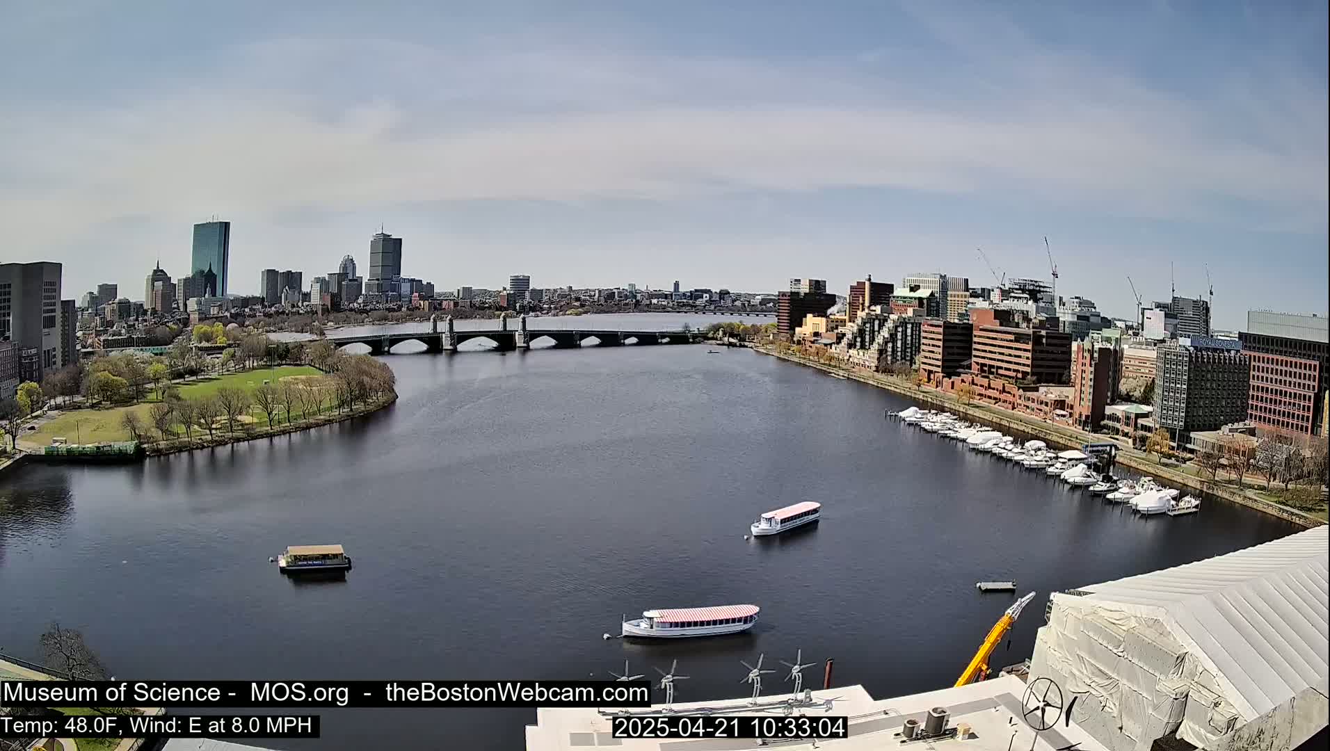 A river flows under a bridge, past a park, and alongside a city skyline on a partly sunny day.