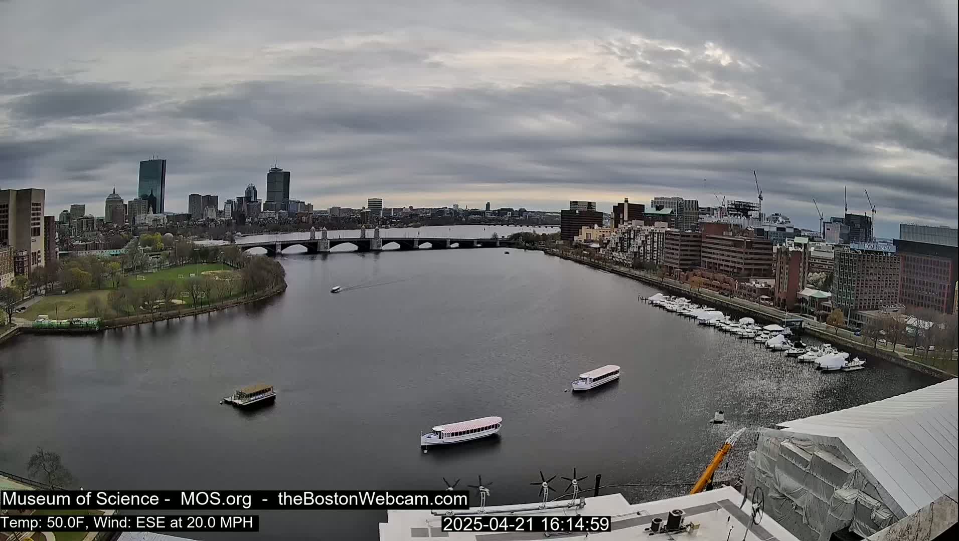A city skyline with a bridge and river is visible under an overcast sky, with several boats on the water and a wind of 20 mph.