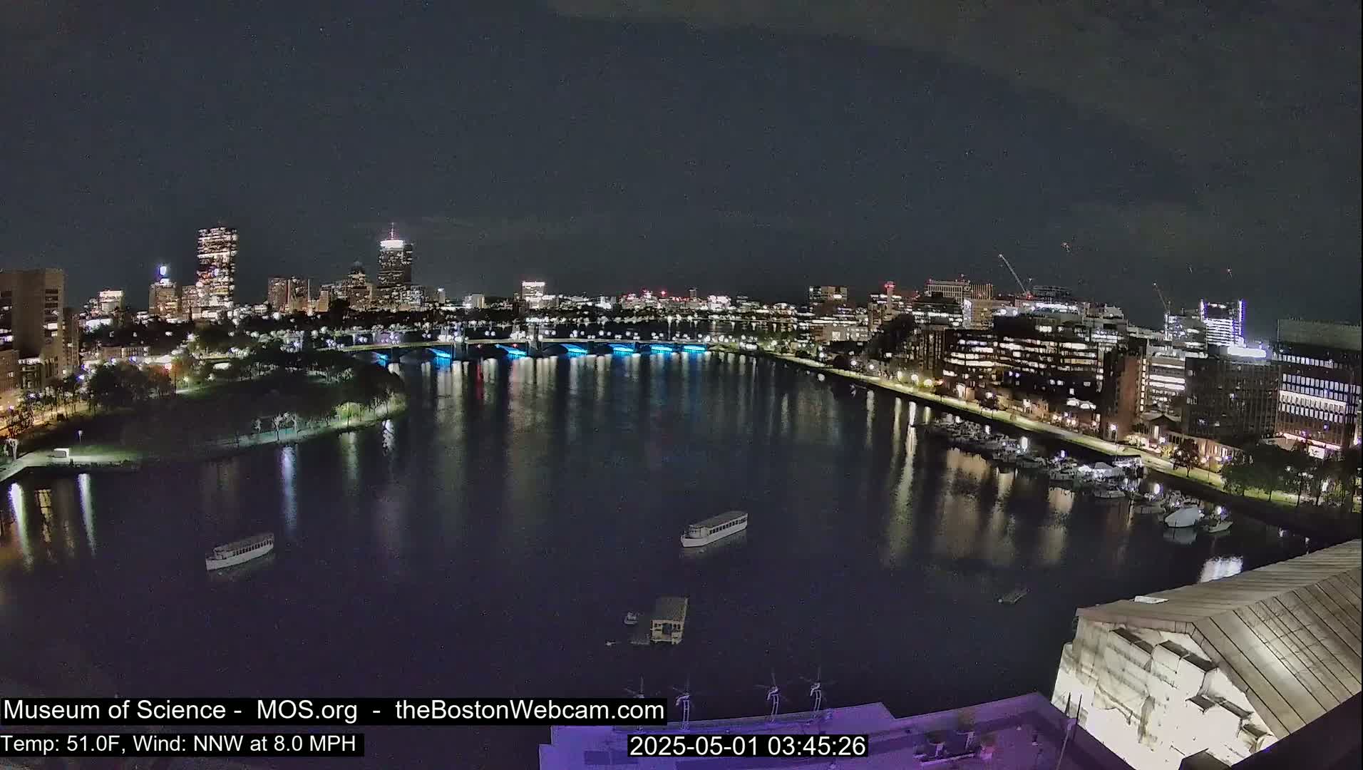 A nighttime view of a city skyline across a dark, calm river, with boats moored along the shore under a partly cloudy sky.