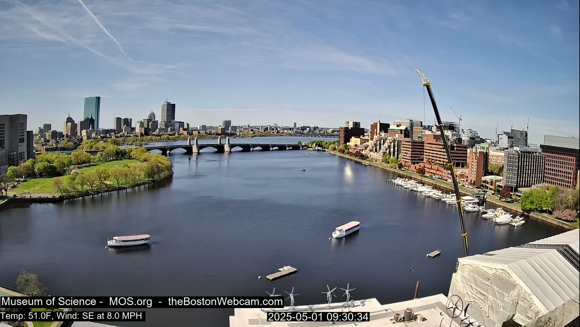 A sunny cityscape is visible across a wide river, with boats on the water and a crane in the foreground.
