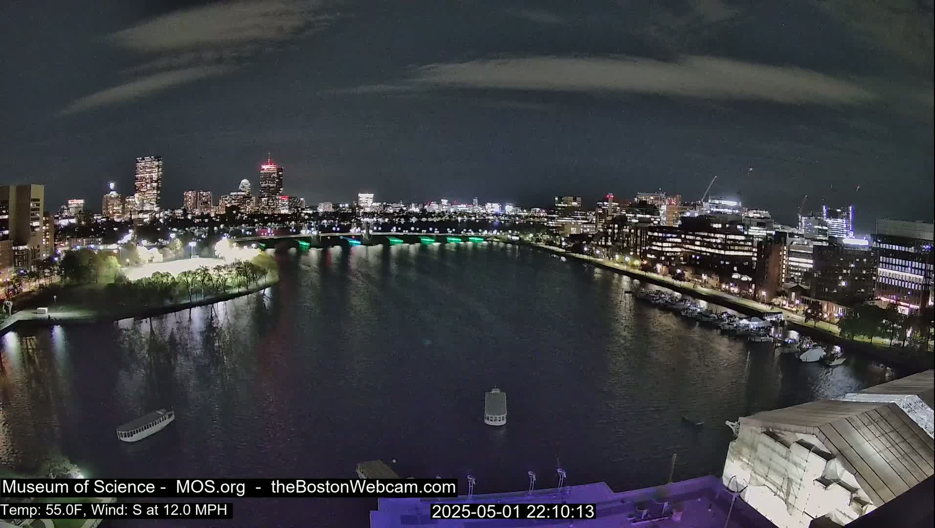 A nighttime view of a city skyline across a calm river, with a few boats moored on the opposite bank under a partly cloudy sky.