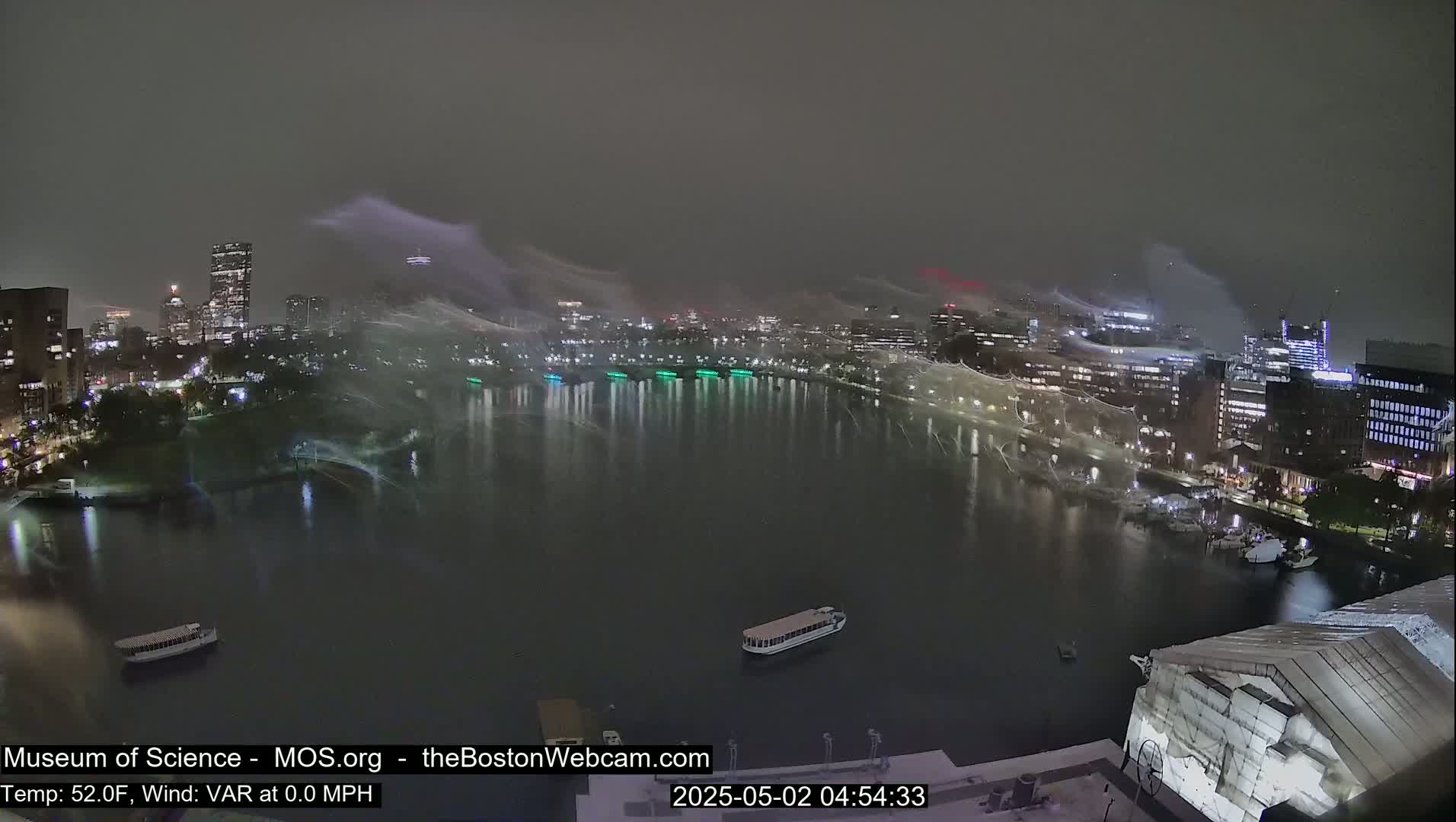 A nighttime cityscape with a body of water in the foreground is shrouded in a light mist or snowfall under a dark, overcast sky.