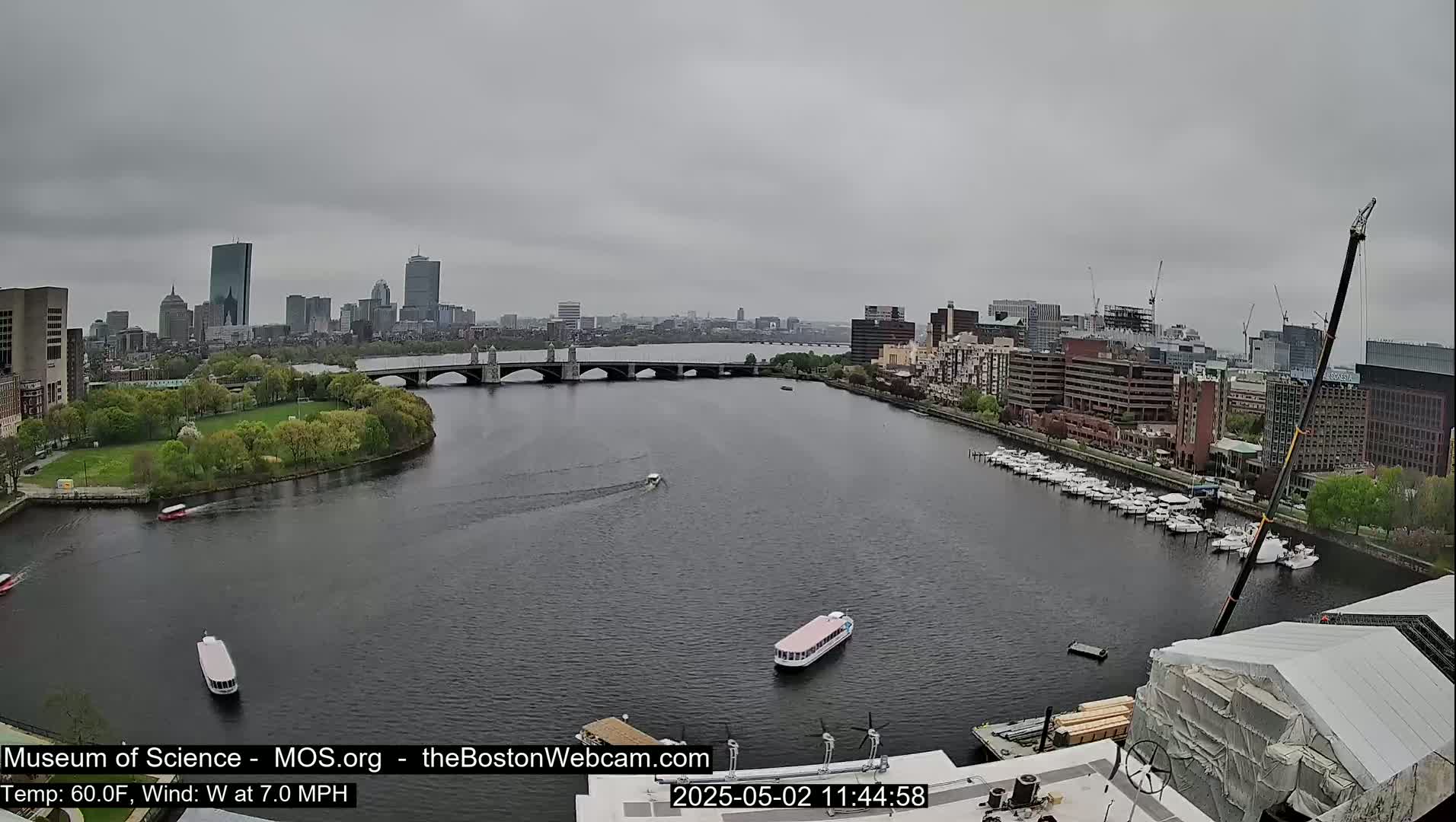 A wide shot of a city skyline across a river on an overcast day, with several boats on the water and a bridge in the distance.