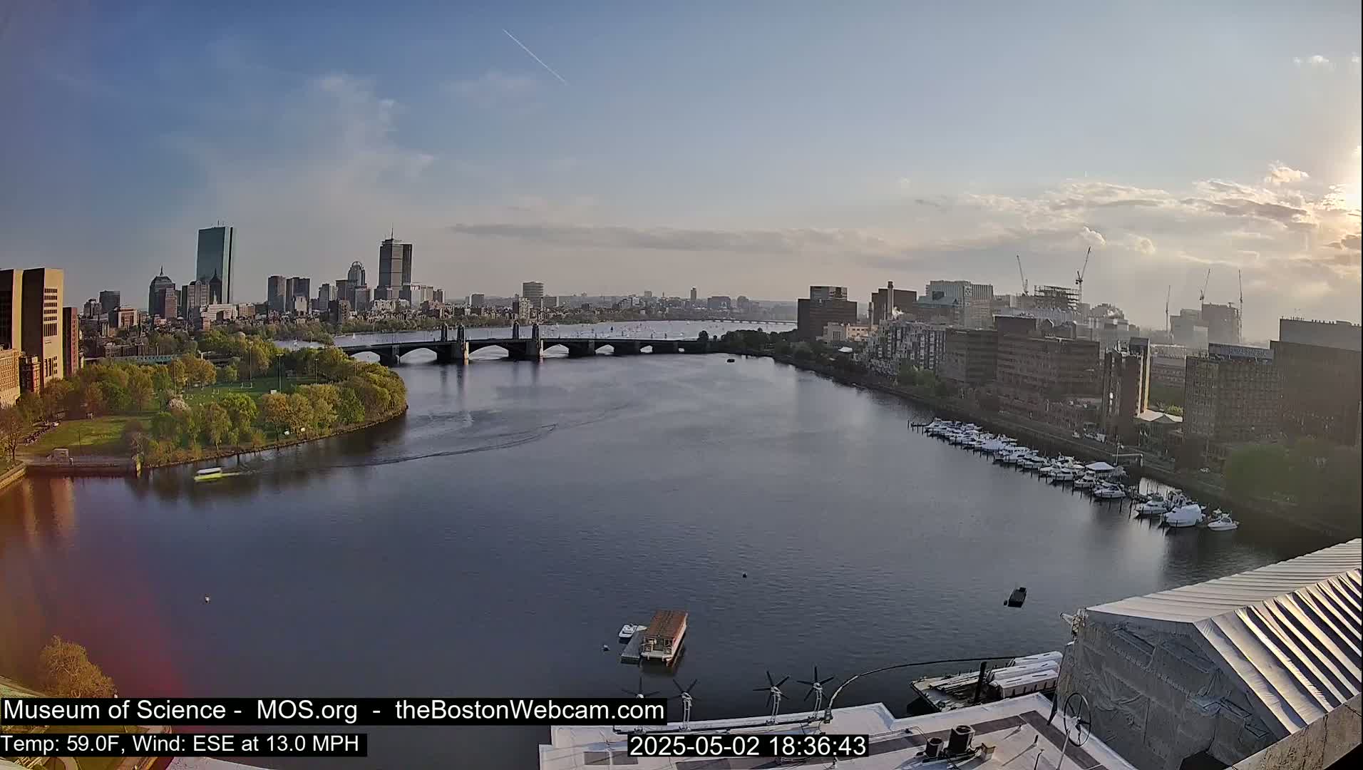 A city skyline with a bridge over a calm river, under a partly cloudy sky.