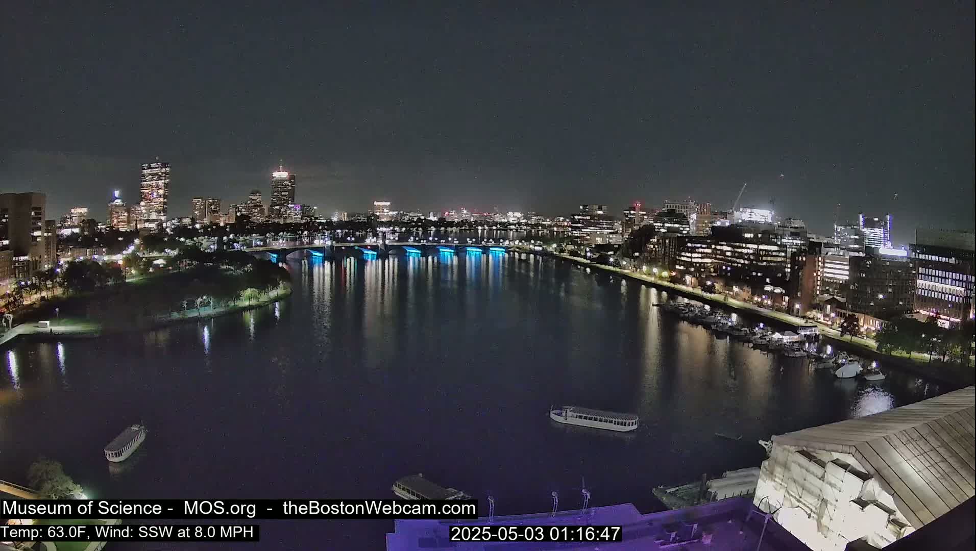 A nighttime view of a city skyline across a calm river, with a bridge lit in blue.