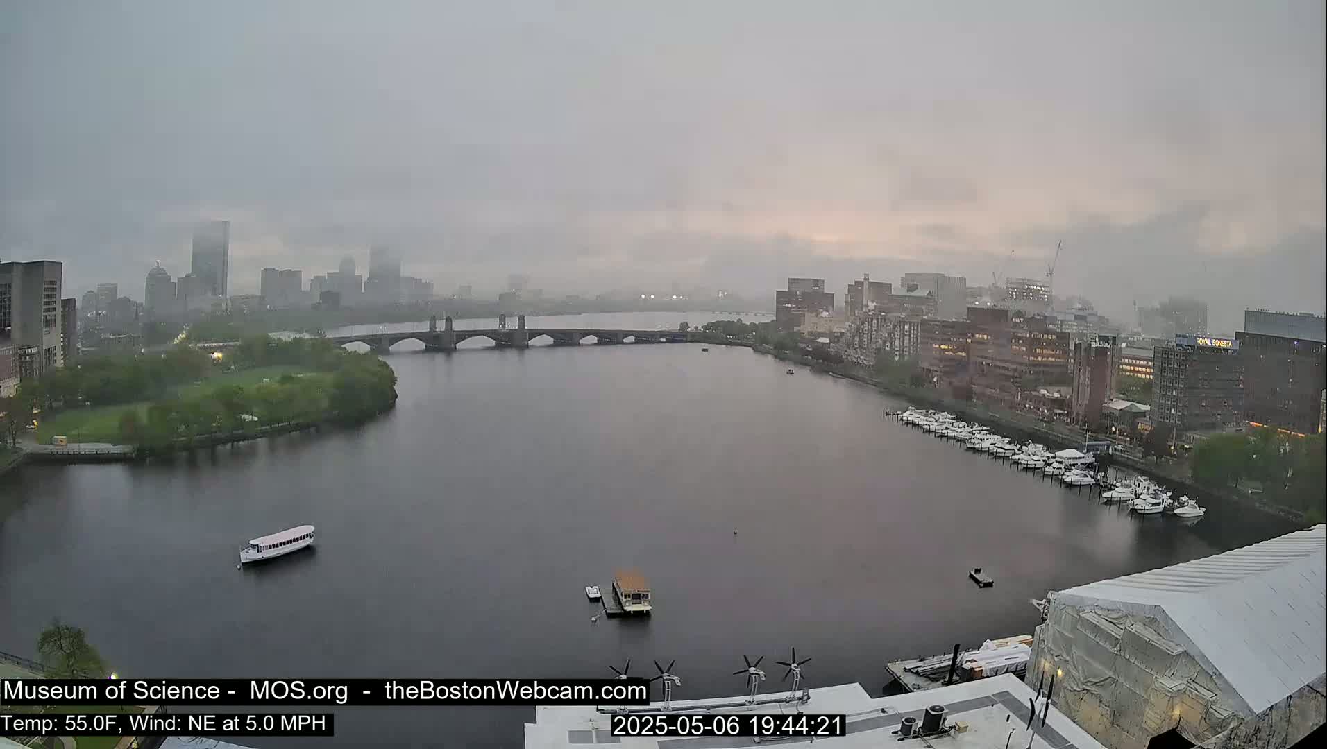A hazy, rainy view shows a river with boats and a bridge leading to a city skyline.