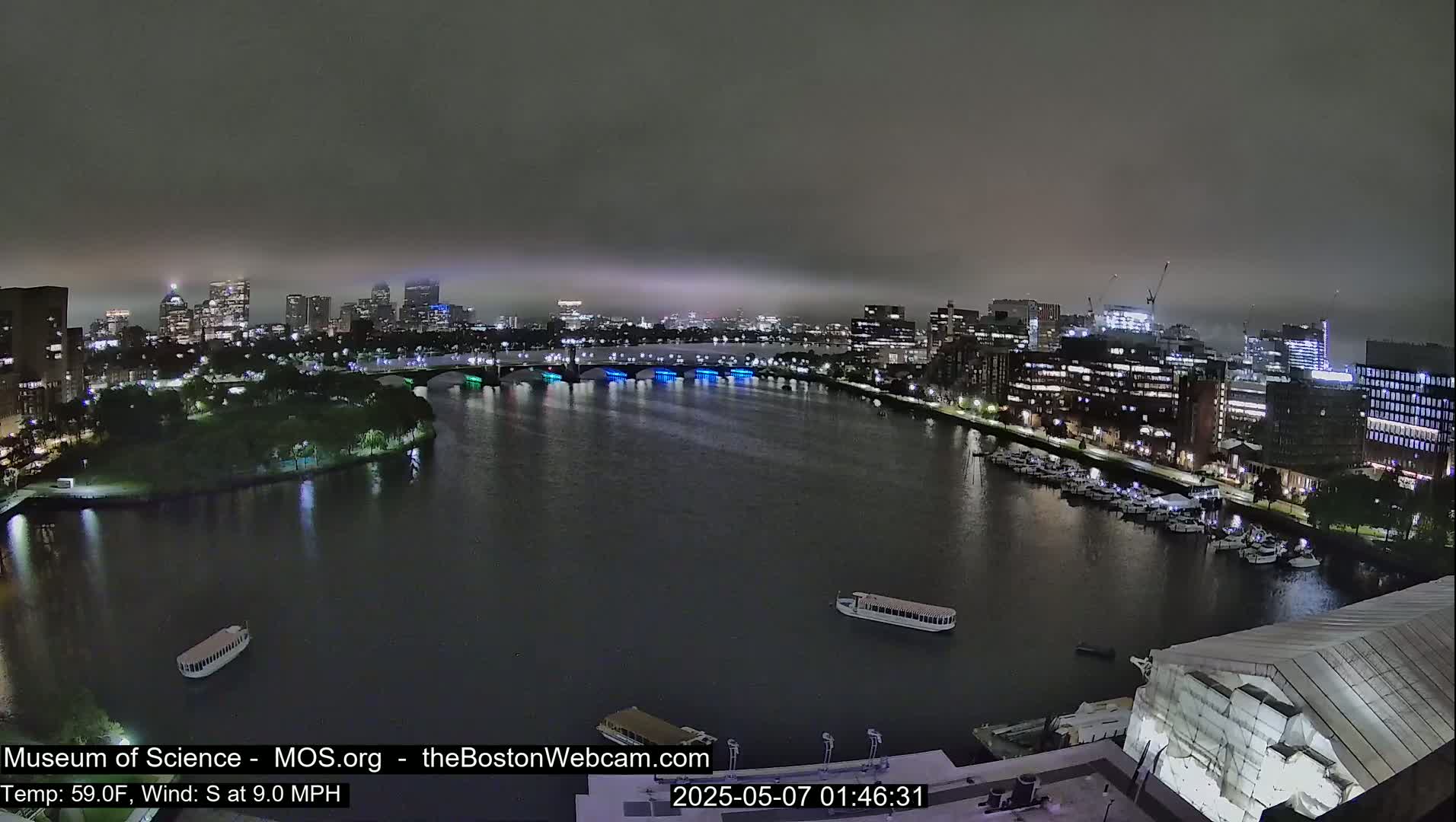 A nighttime, foggy view of a city skyline across a river, with boats docked along the waterfront and a bridge lit with colorful lights.