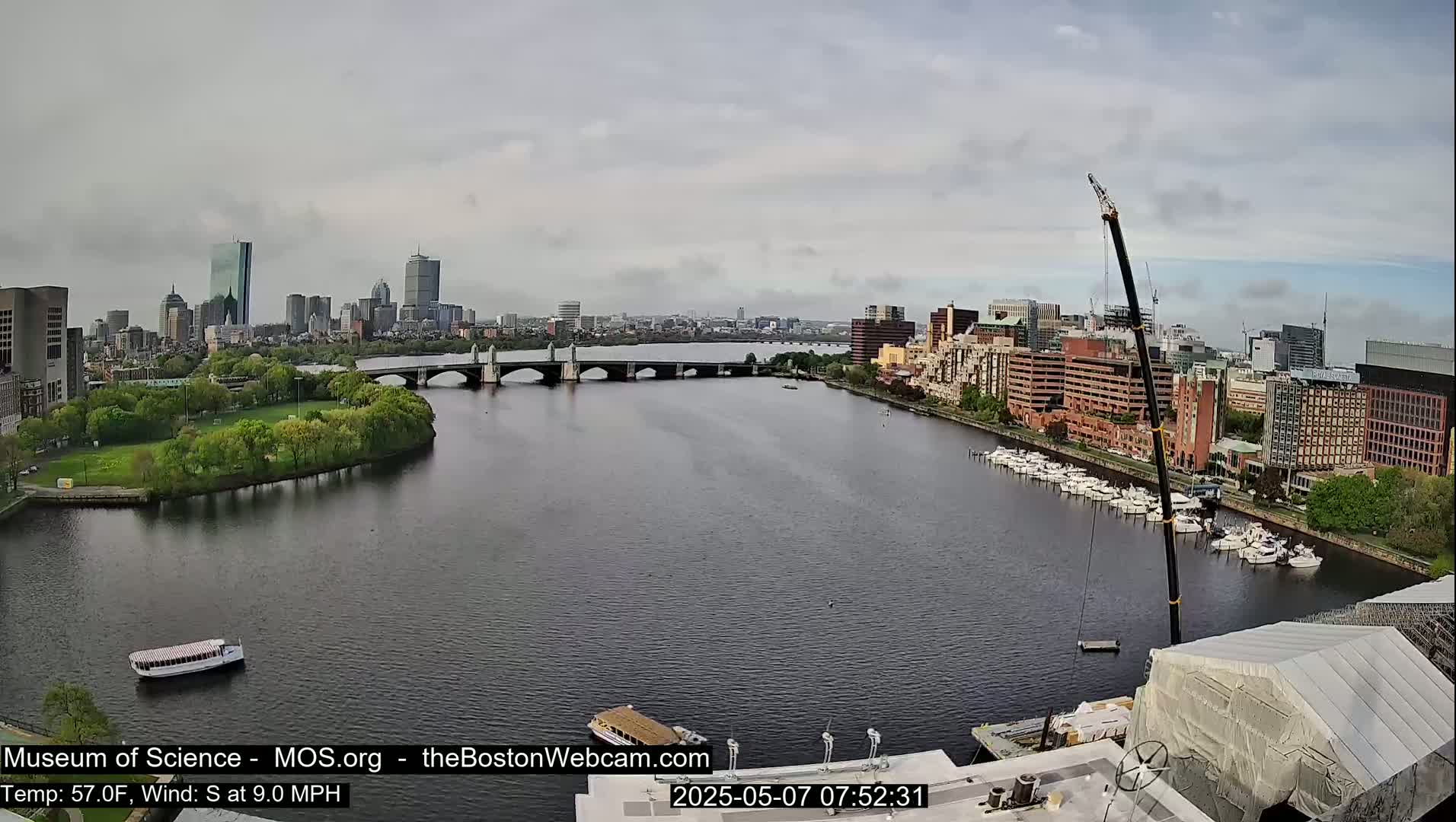 A city skyline with a bridge over a river, under partly cloudy skies, with a boat and a crane visible.