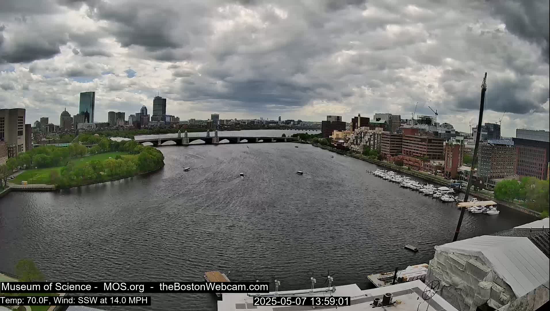 A wide shot of a river with a bridge, boats, and a city skyline under a cloudy sky.