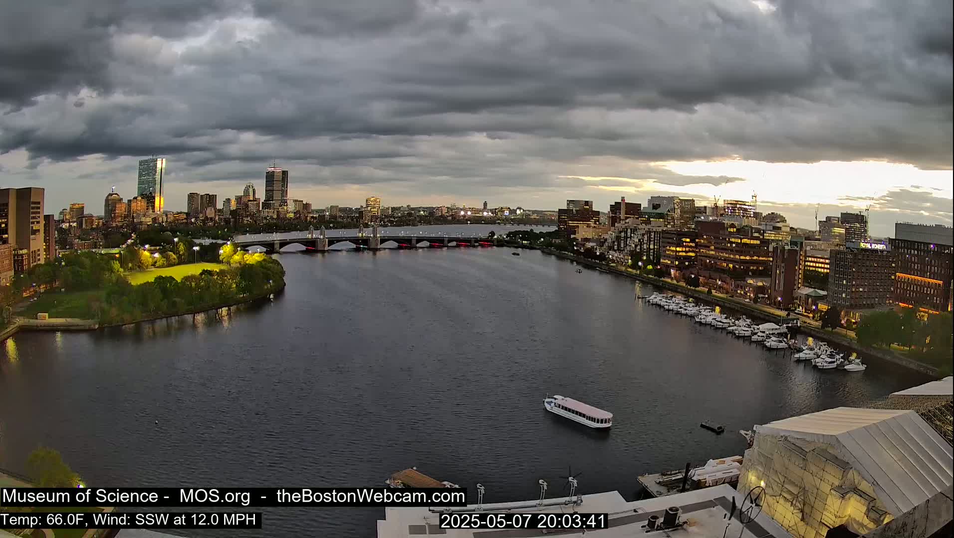 A city skyline at dusk under a cloudy sky, viewed from across a body of water with a bridge and docked boats.
