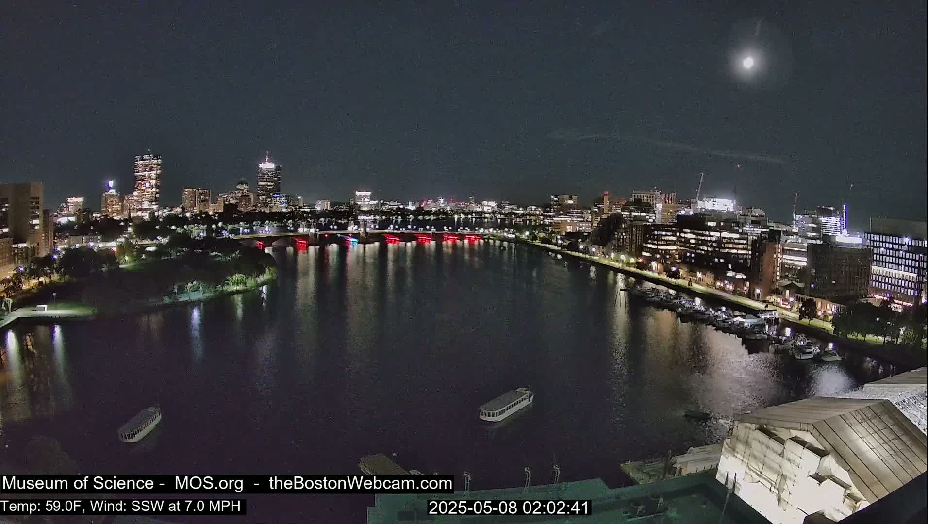A nighttime cityscape with a river reflecting city lights, a few boats on the water, and a bright moon in the sky under clear conditions.