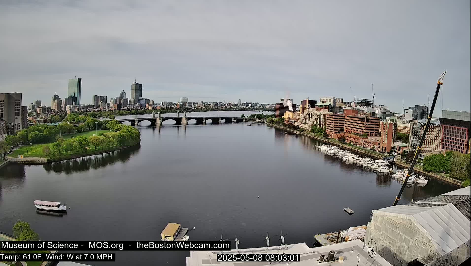 A river flows past a city skyline under a cloudy sky, with a bridge, boats, and a construction crane visible.