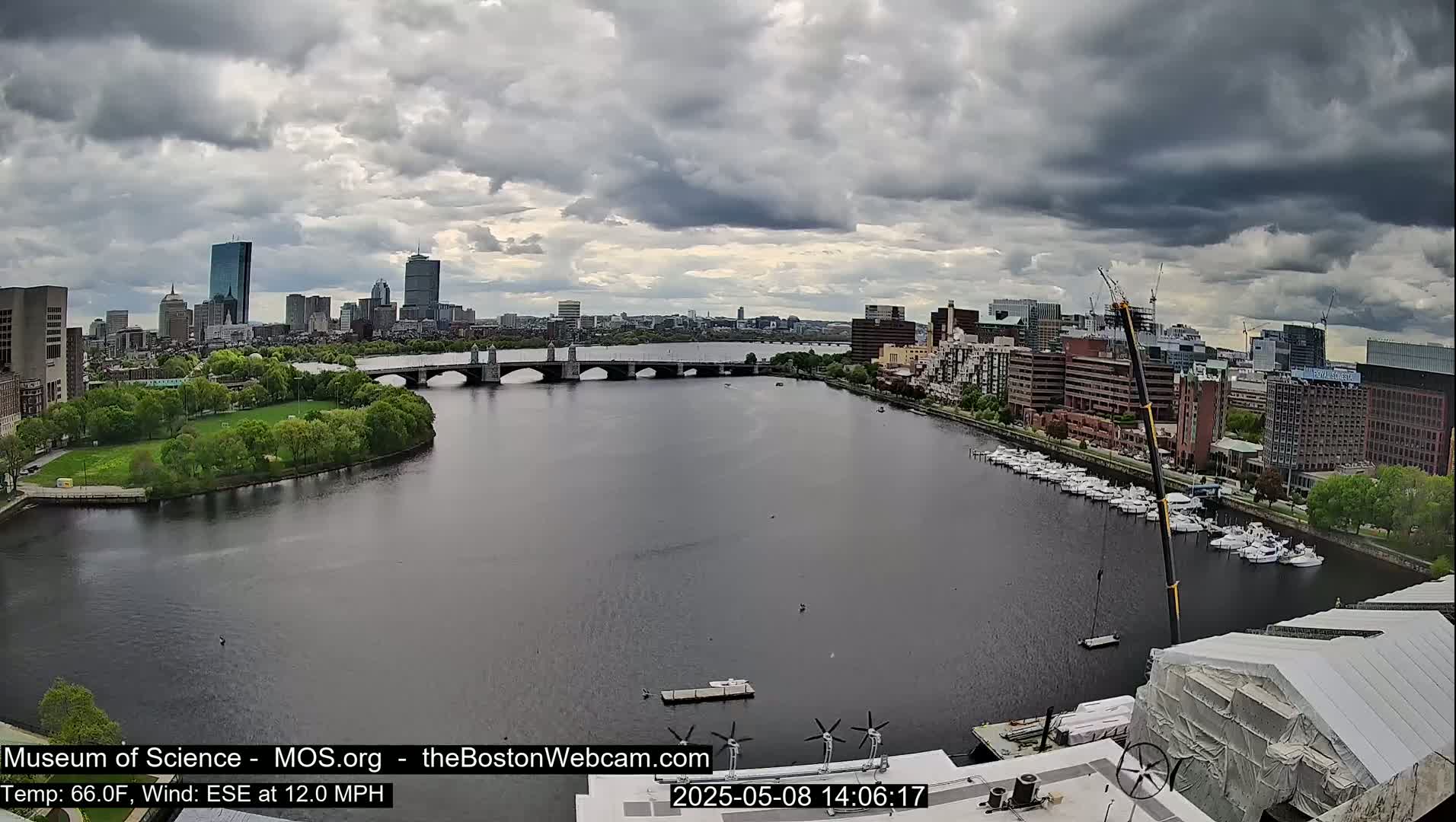 A city skyline is visible across a wide river under a cloudy sky, with a bridge spanning the waterway and numerous boats docked along the shore.