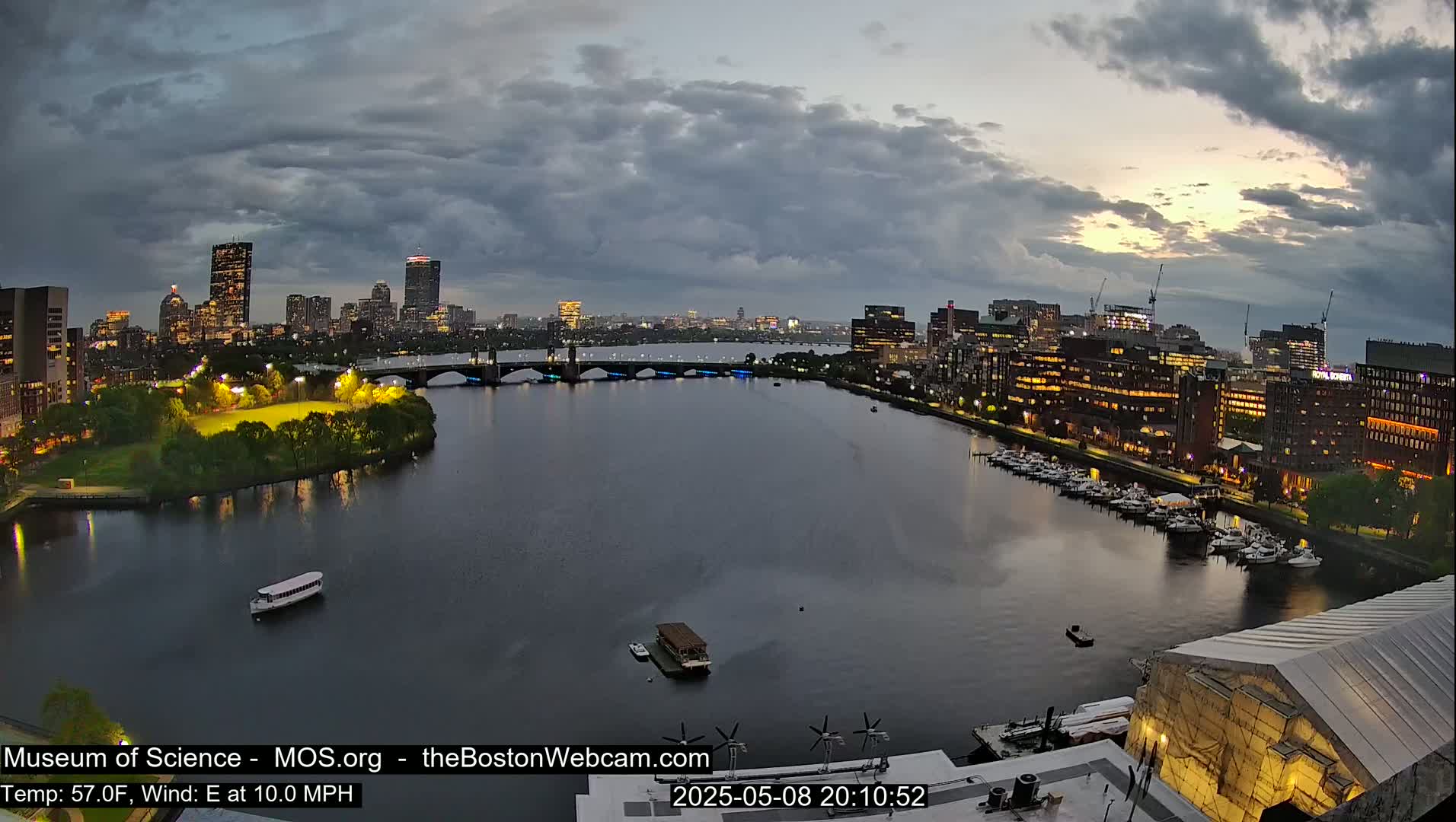 A nighttime view of a city skyline across a calm river under a cloudy sky, with boats on the water and a lit-up park along the shore.