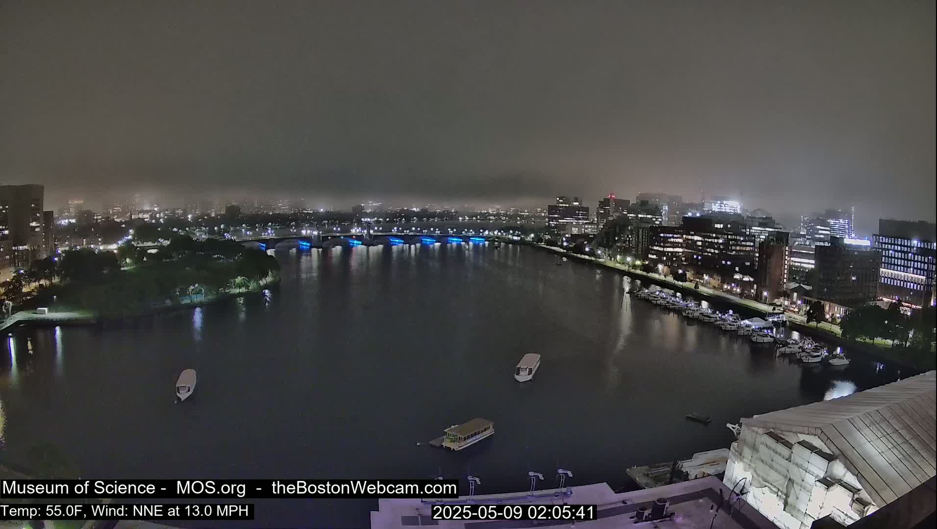 A nighttime view of a city waterfront with a river, bridges, buildings, boats, and a hazy atmosphere with a 13 mph wind.