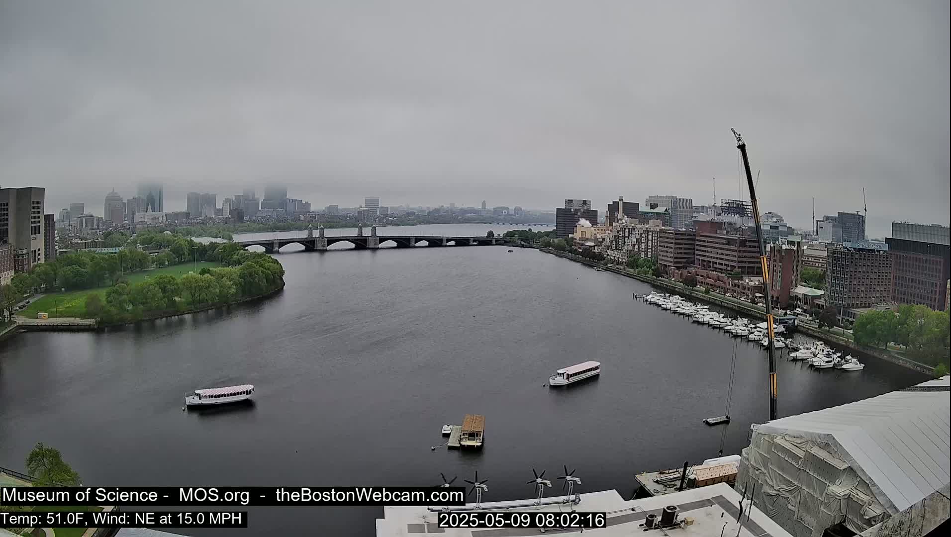 A hazy, overcast day shows a city skyline across a river with a bridge, boats, and a construction crane.