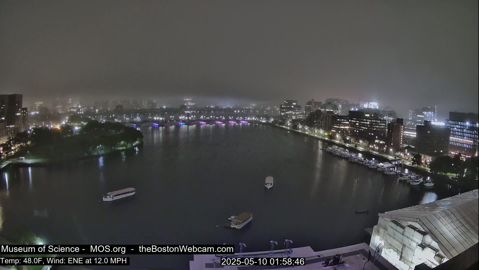 A nighttime view of a city waterfront shrouded in fog and mist, with boats on the water and city lights reflecting on the dark water.
