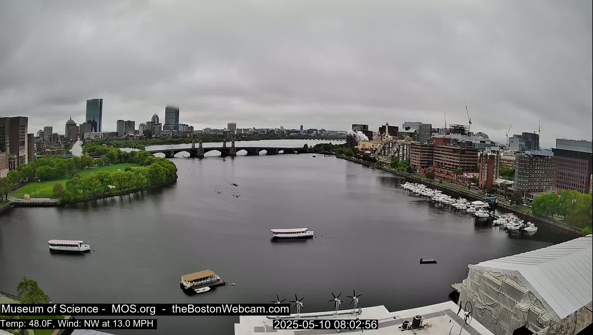 A river with several boats and a bridge is seen under an overcast sky, with a city skyline in the background.