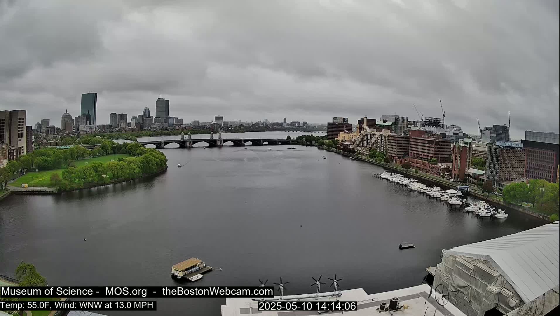 A city skyline is visible across a wide river under an overcast sky, with a bridge, docked boats, and green parkland.
