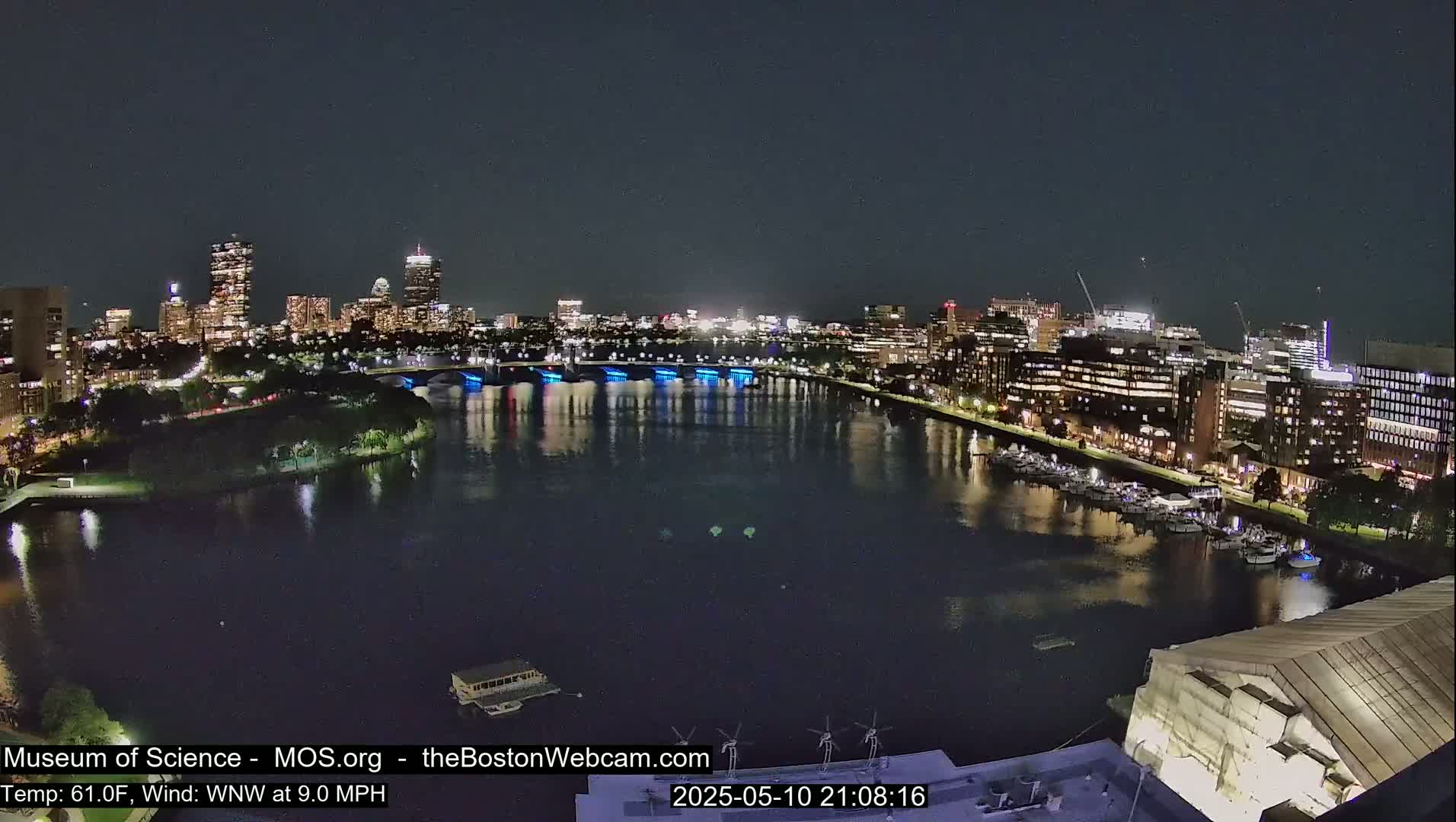 A nighttime view of a city skyline across a calm river, with a bridge illuminated in blue.