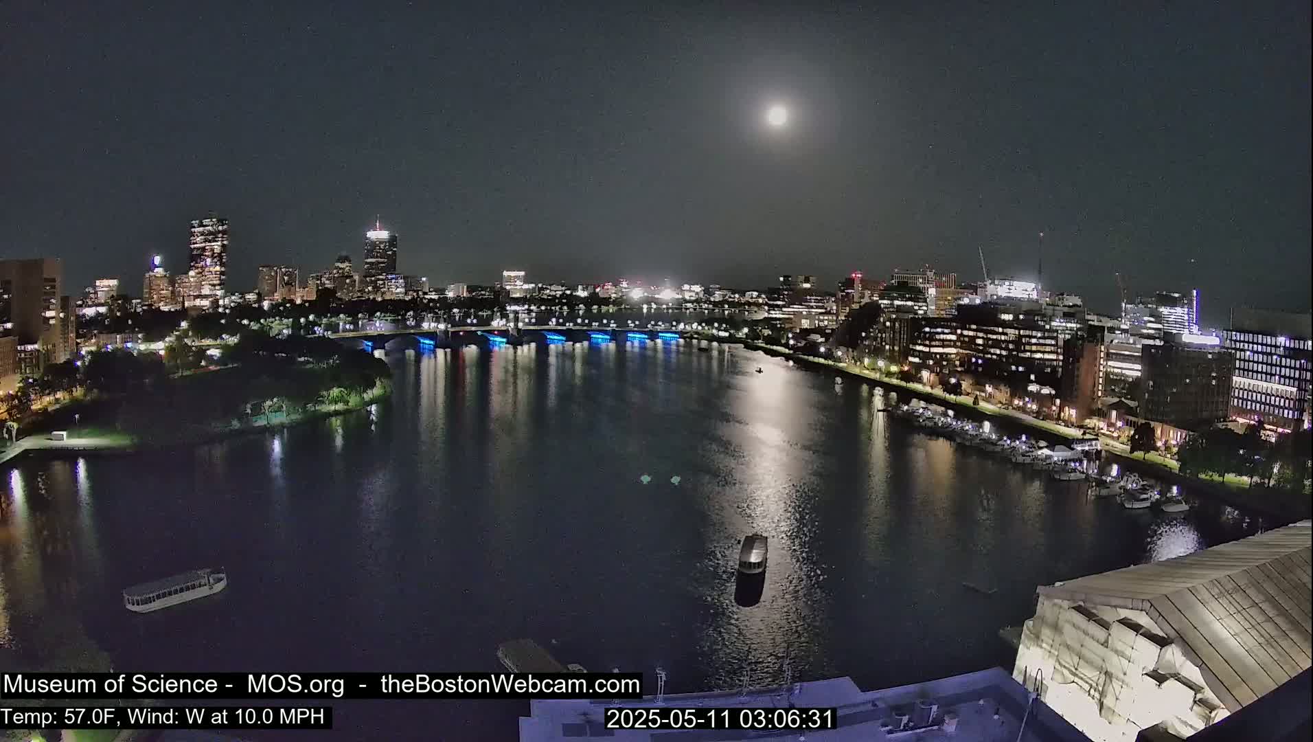 A nighttime cityscape with a bright moon, a river reflecting city lights, and boats docked along the shore under a clear sky.