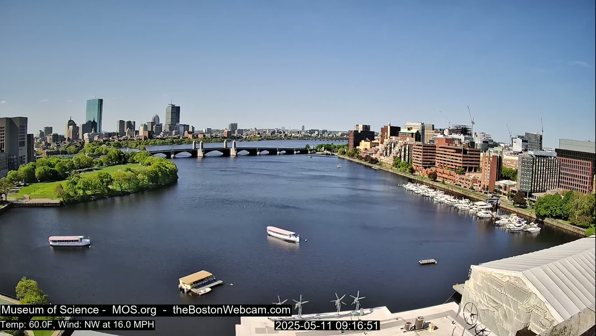 A city skyline and river are visible on a sunny day with a northwest wind of 16 mph and a temperature of 60°F; several boats are on the water and docked along the shore.