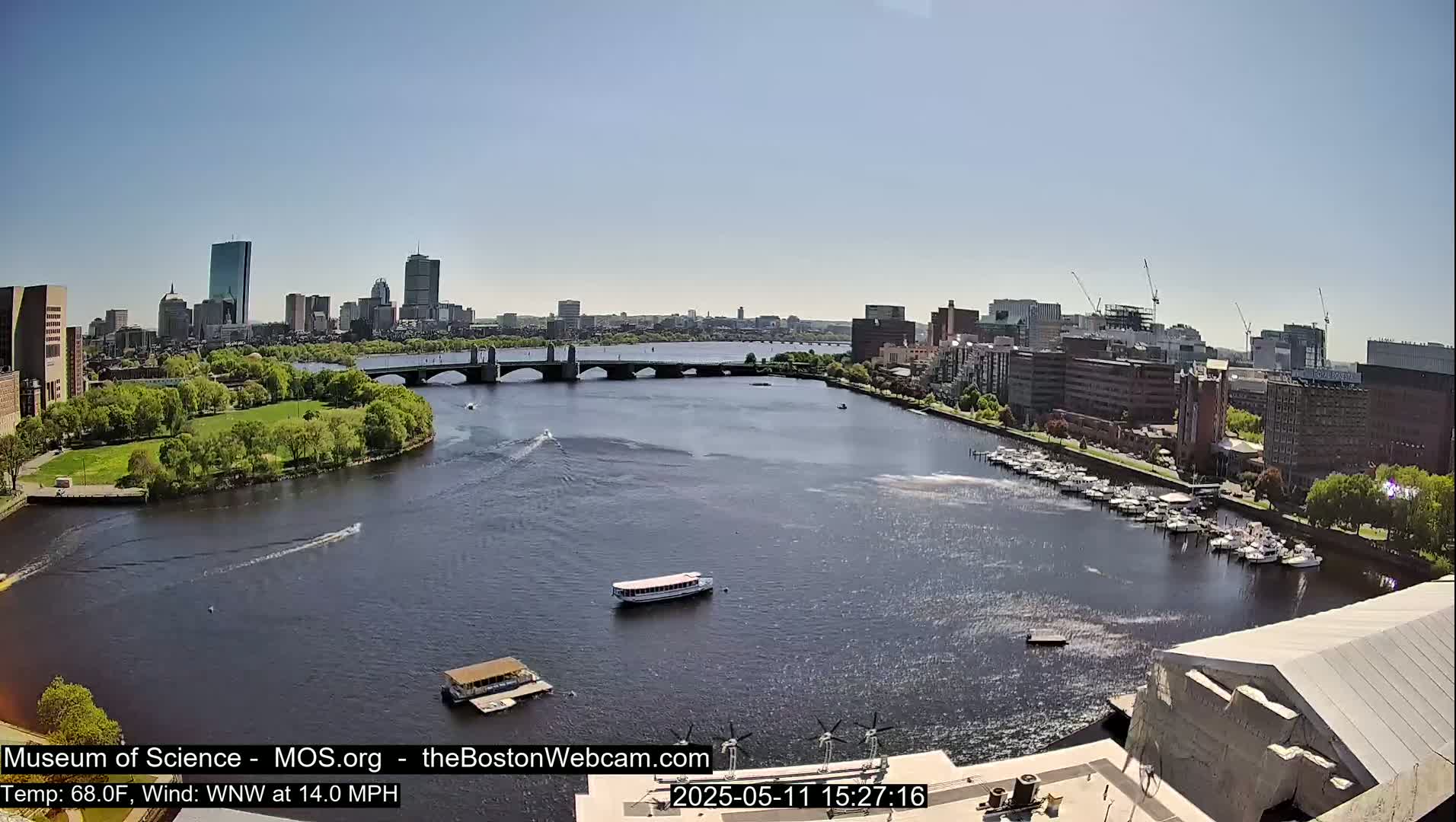 A sunny day shows a city skyline across a river with a bridge, boats, and a marina;  a large building is partially visible in the foreground.