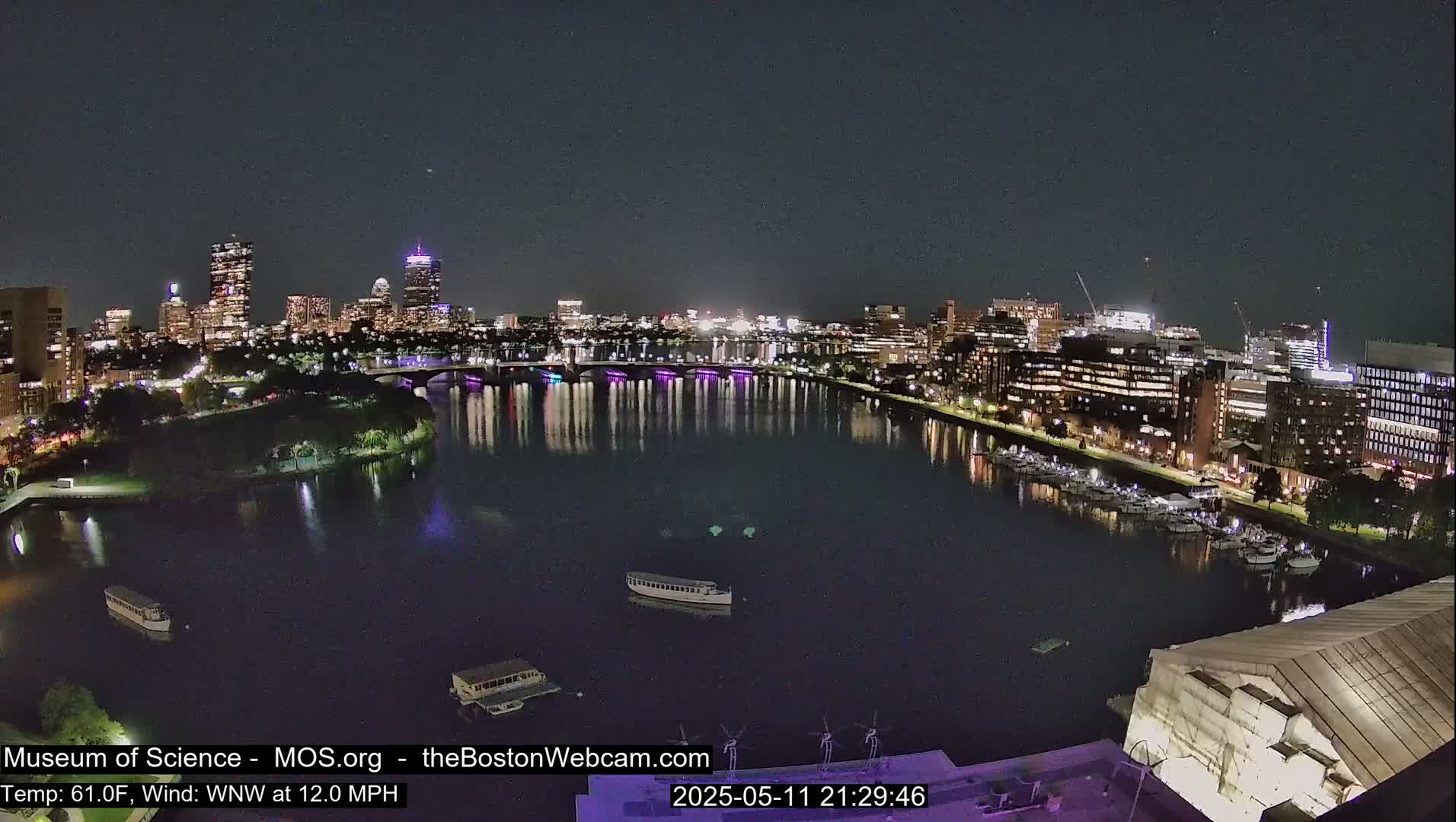 A nighttime view of a city skyline across a calm river, with boats moored along the waterfront under a clear sky.