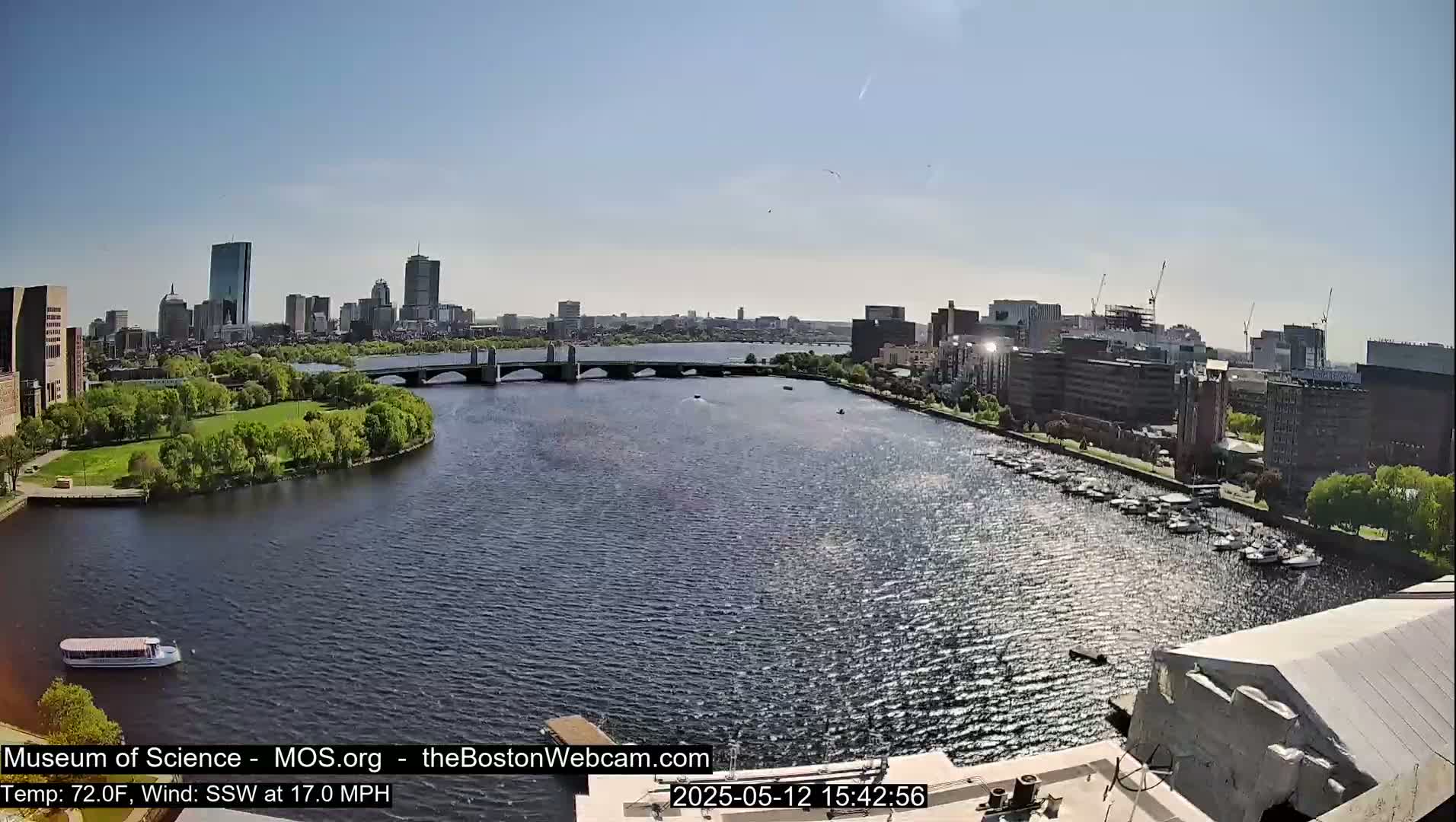 A sunny cityscape is shown across a body of water, with a bridge and several boats visible, under a clear sky.