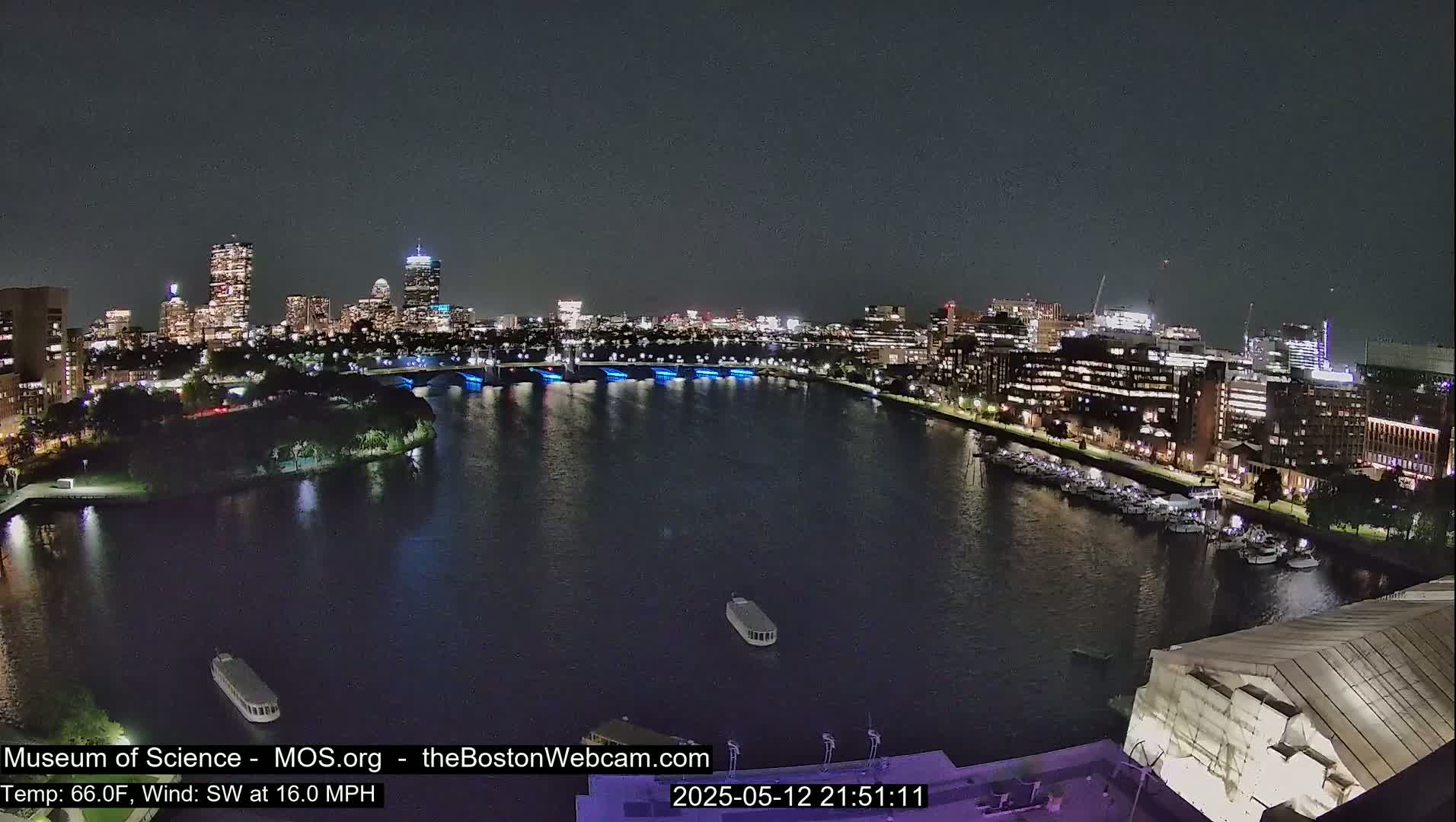 A nighttime view of a city skyline across a dark river, with boats on the water and a southwest wind at 16 mph.