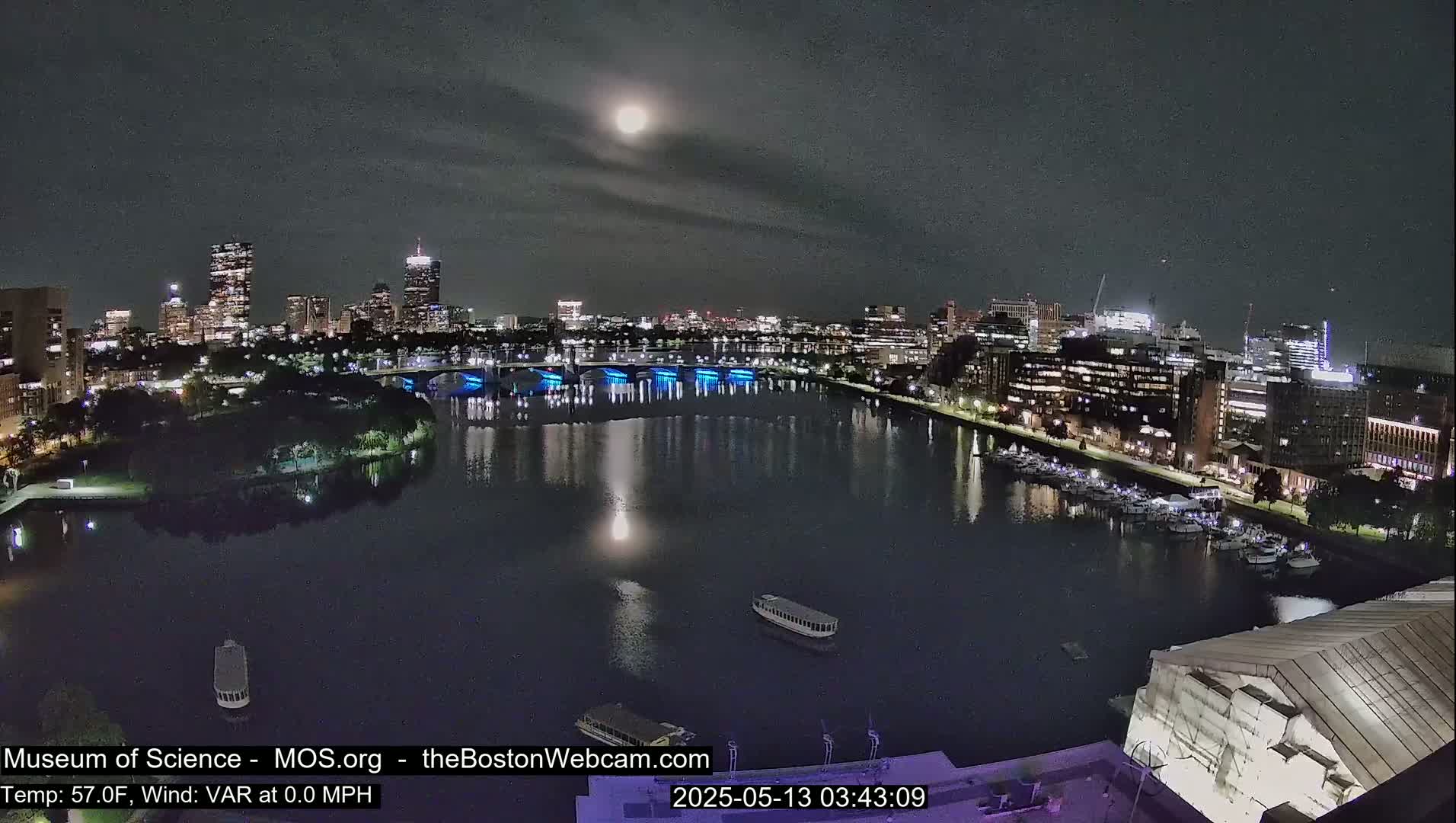 A nighttime view of a city skyline across a calm river under a bright moon and mostly clear sky.