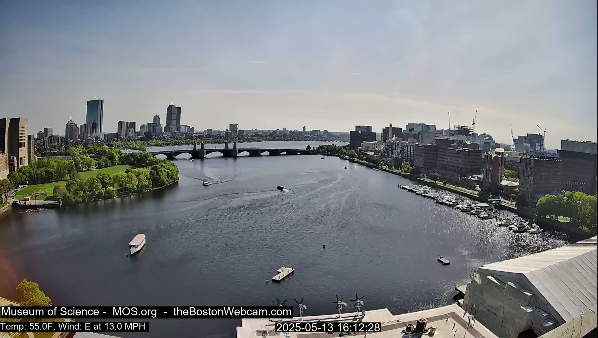 A sunny cityscape with a river and bridge, showing boats on the water and a marina with docked vessels.