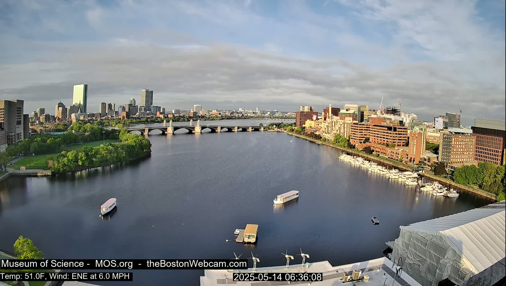 A city skyline is visible across a calm river with several boats and a bridge under partly cloudy skies.