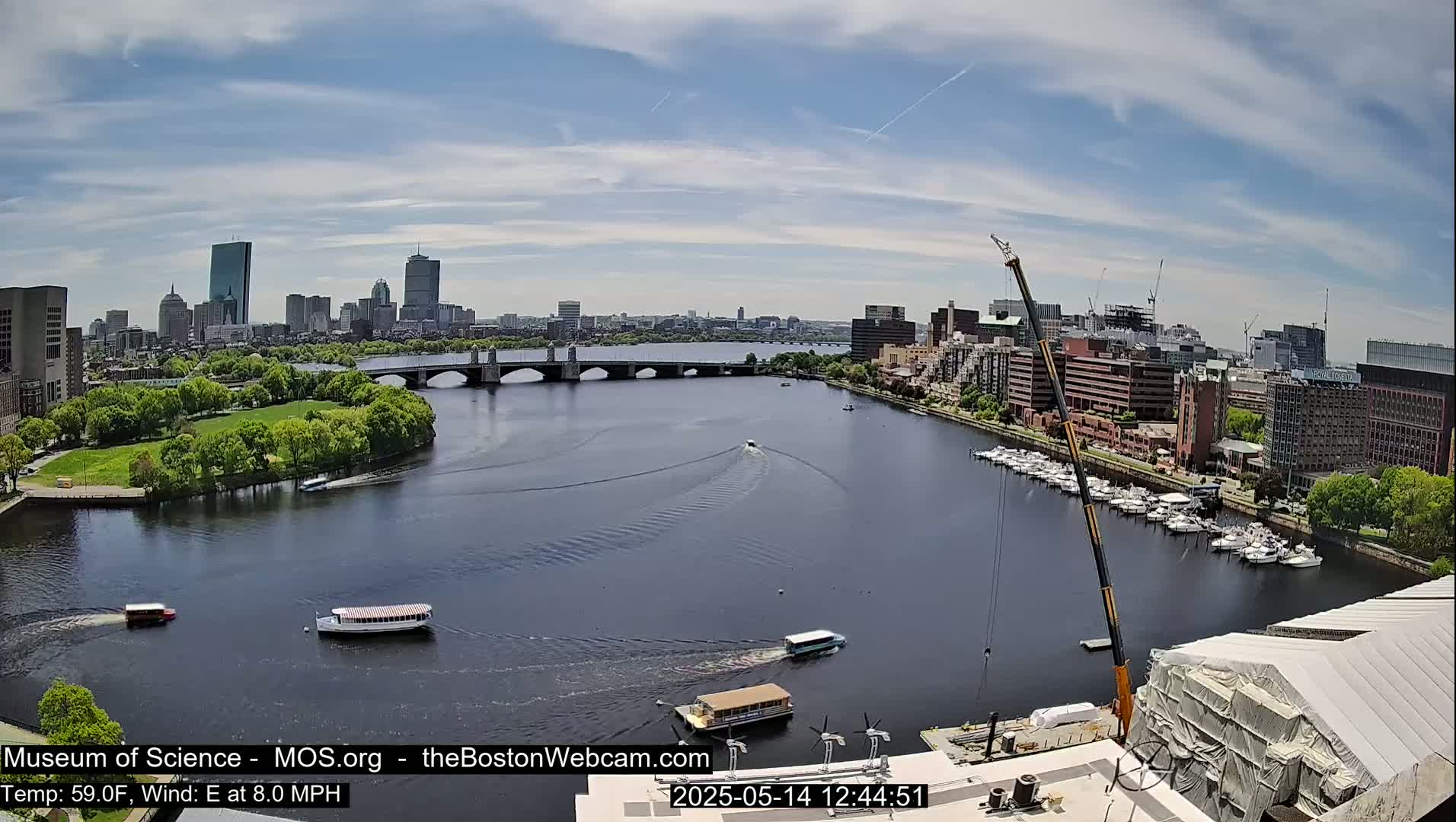 A partly cloudy day shows a city skyline across a river with several boats and a bridge, a crane, and a marina.