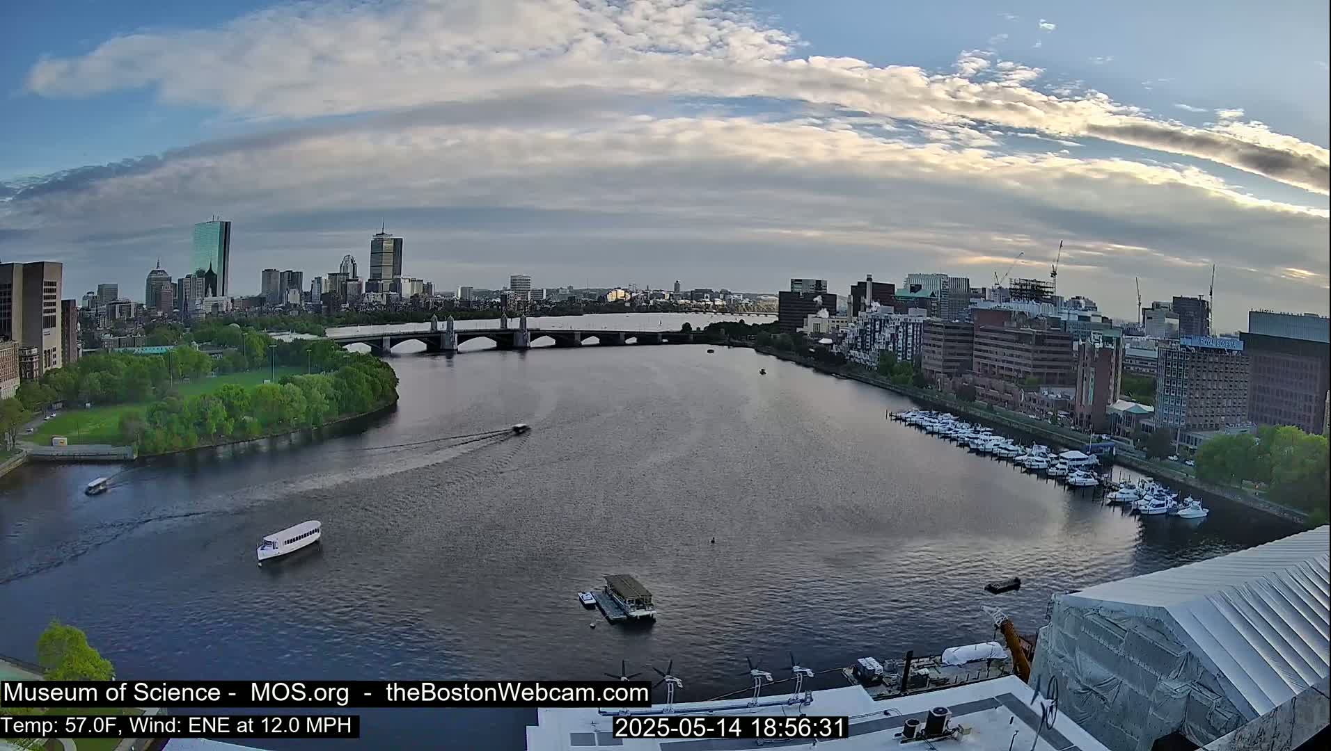 A partly cloudy evening scene shows a river with boats and a bridge leading to a city skyline.