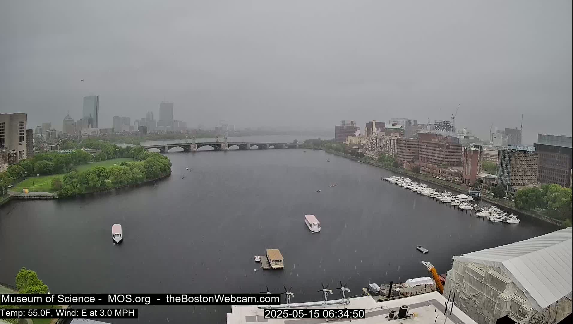 A rainy view of a river with boats and a bridge, showing a city skyline in the background.