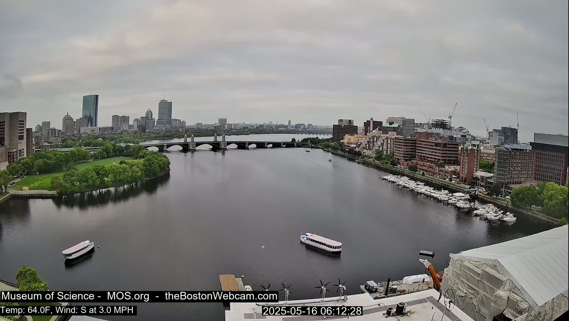 A city skyline is visible across a river on an overcast day, with boats docked along the shoreline and a bridge spanning the waterway.