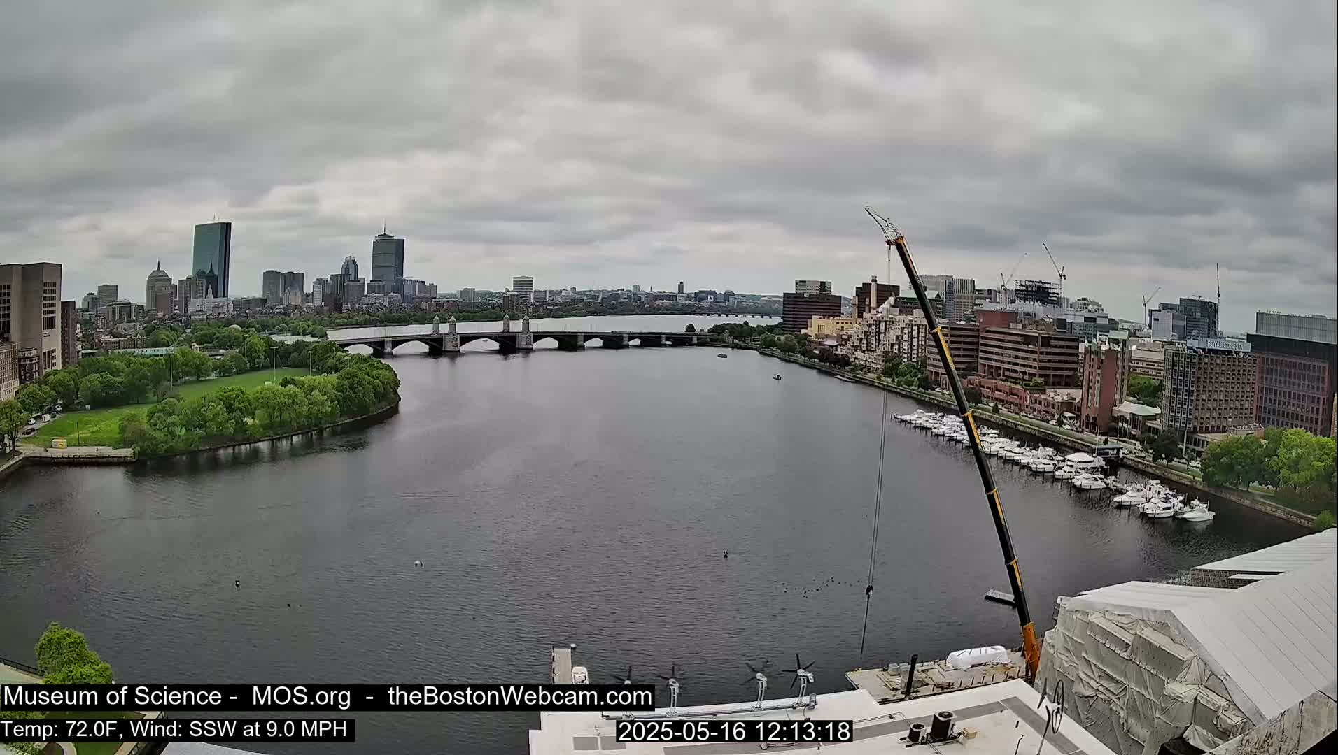 A wide shot of a city skyline across a river on an overcast day, with a large crane and several boats visible.