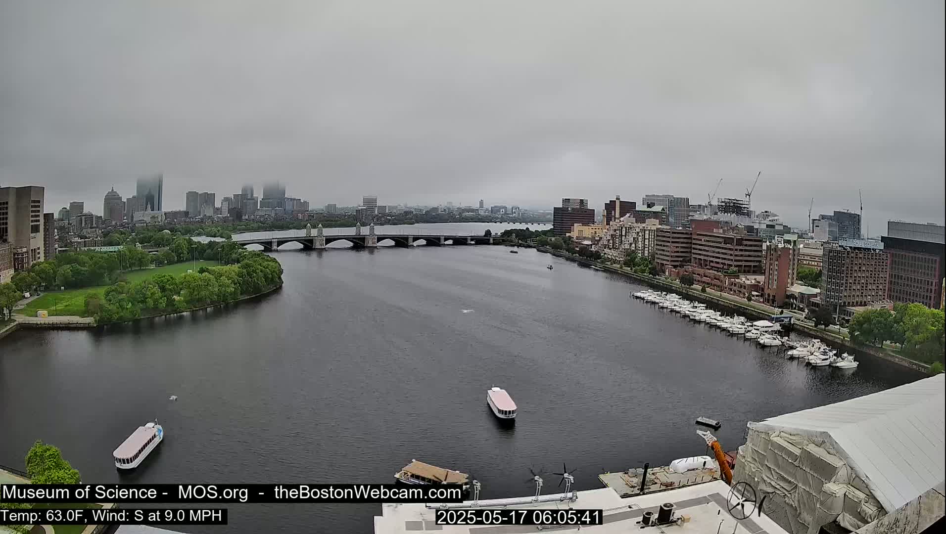 A city skyline is visible across a river on an overcast day, with several boats on the water and a bridge in the midground.