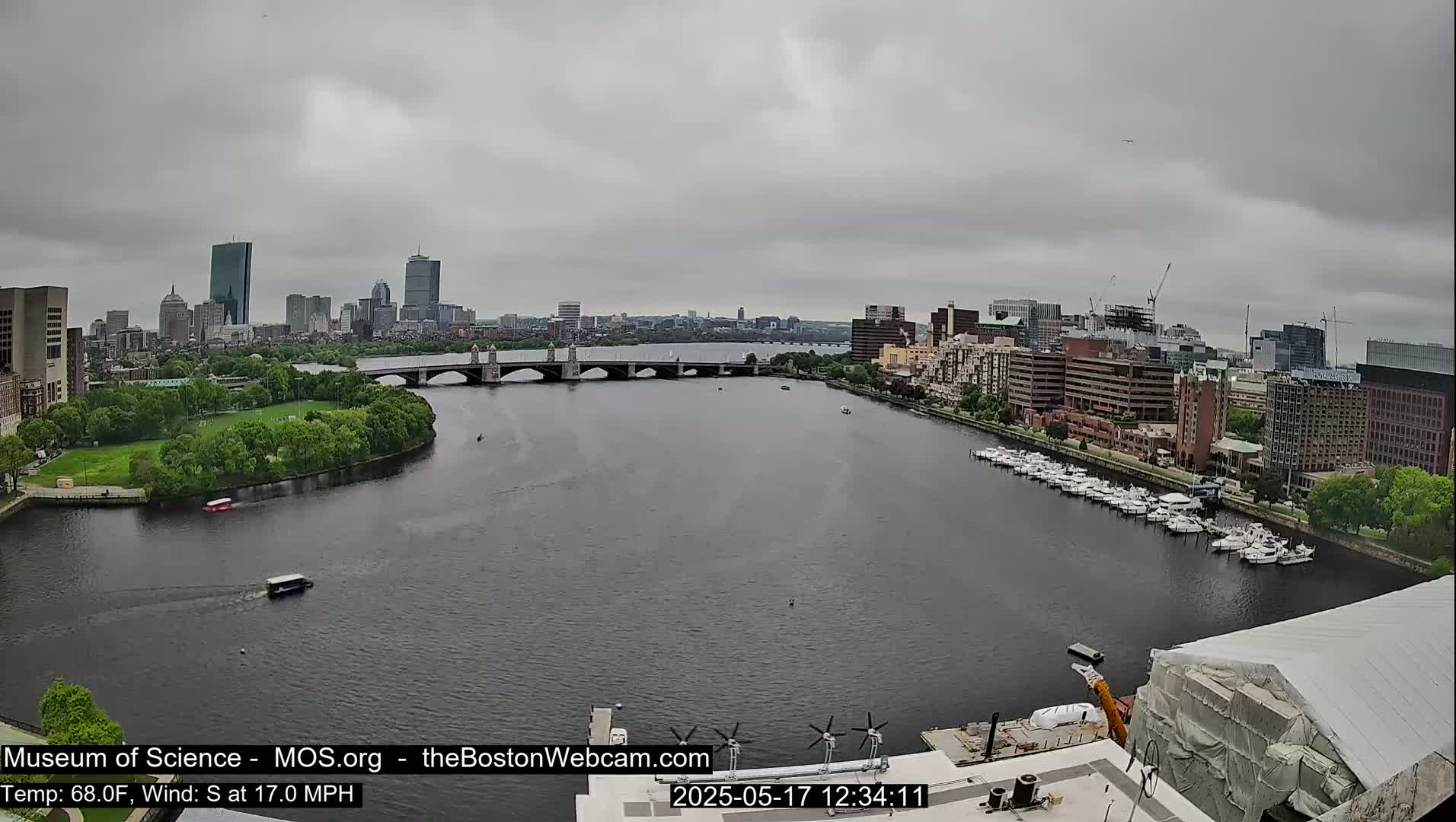 A city skyline is visible across a river on an overcast day, with several boats docked along the shoreline and a bridge connecting the two sides.