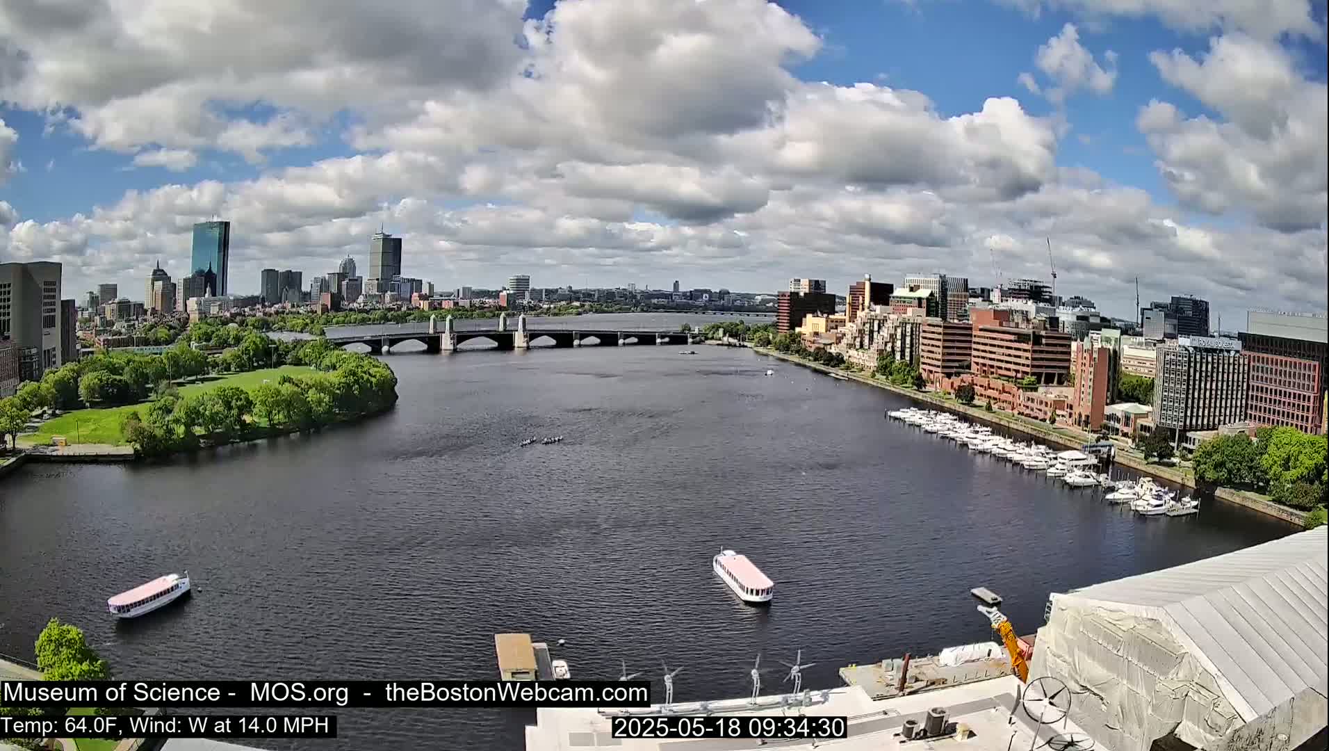 A partly cloudy day shows a city skyline across a wide river, with boats docked along the shore and a bridge spanning the water.
