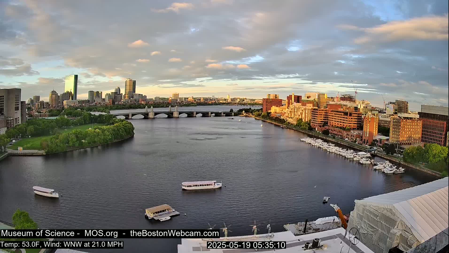 A wide, calm river flows past a city skyline under a partly cloudy sky, with several boats docked along the riverbank and a few more traveling on the water.