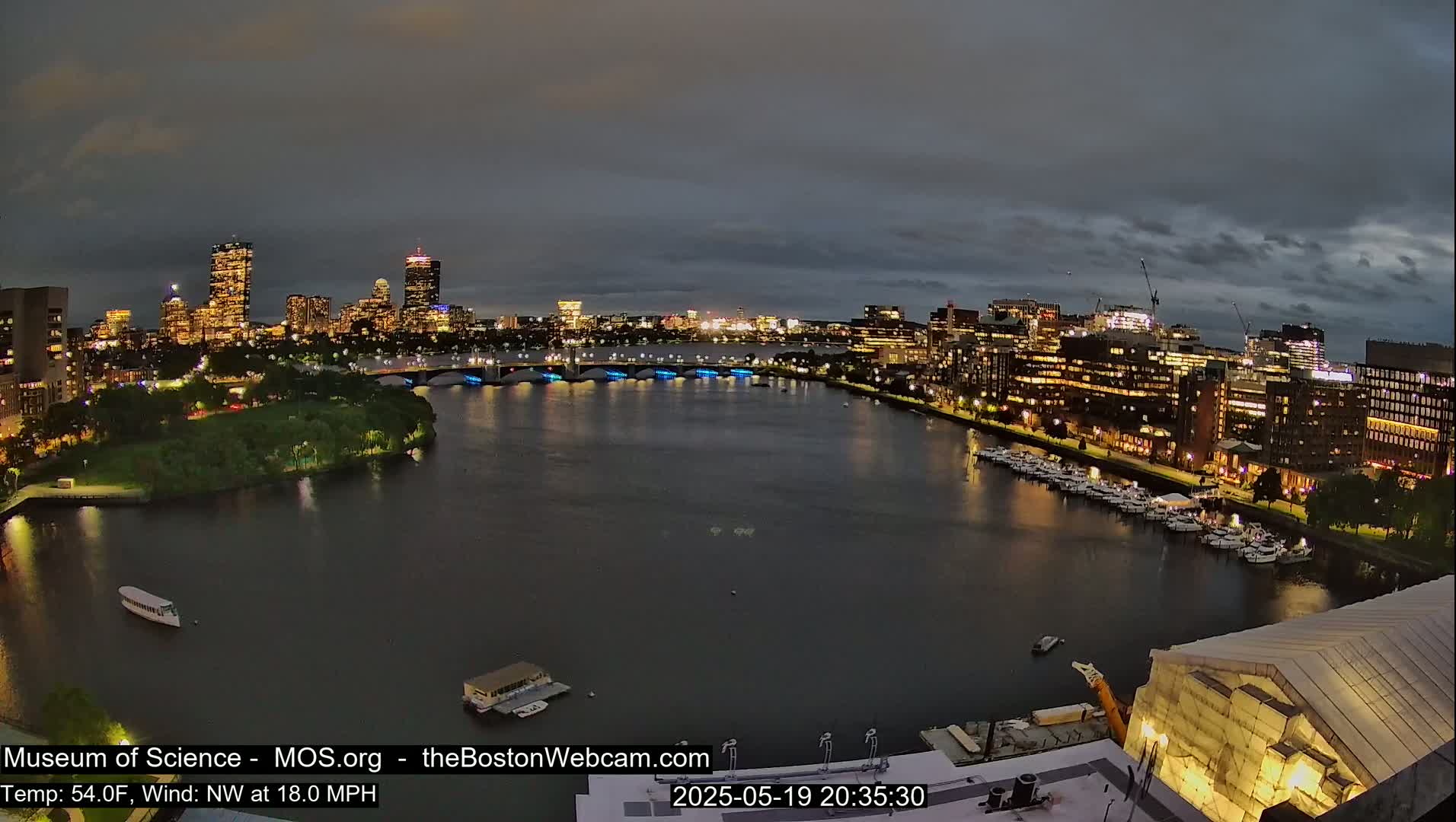 A nighttime view of a city skyline across a river, under a partly cloudy sky, with a temperature of 54°F and a northwest wind at 18 mph.